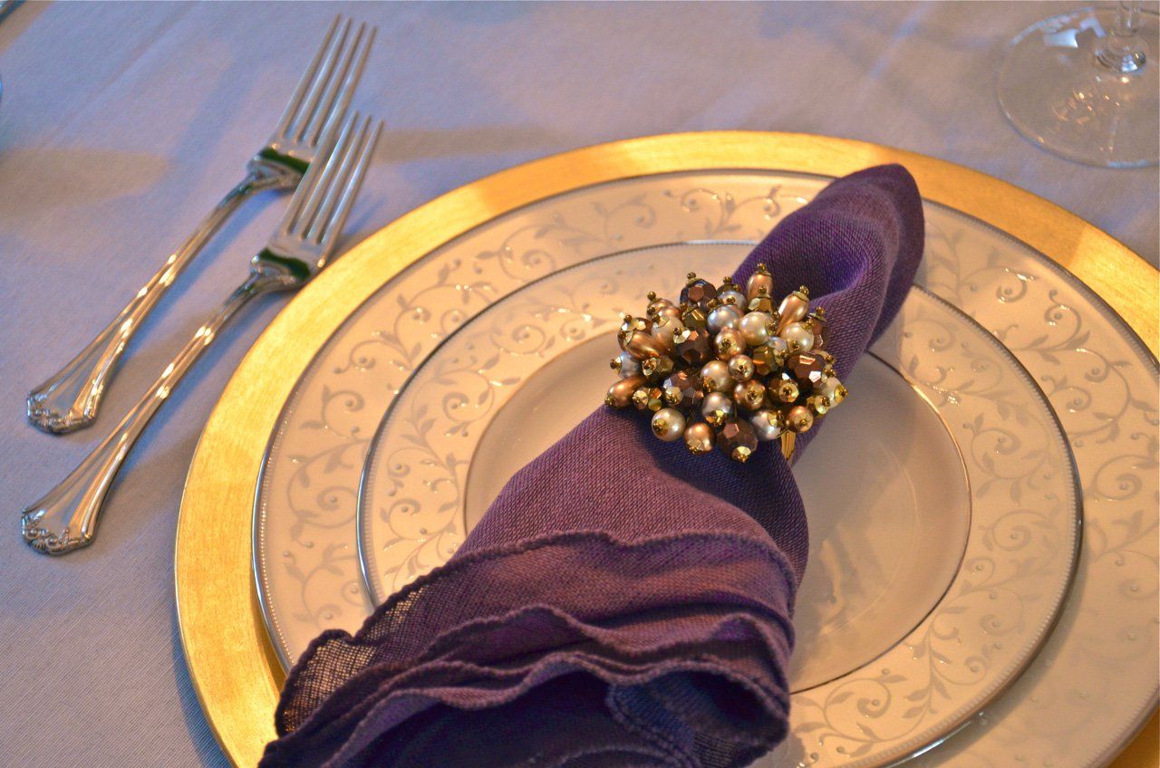Place setting with purple napkin in a beaded ring, gold charger, and two forks on a light blue tablecloth.