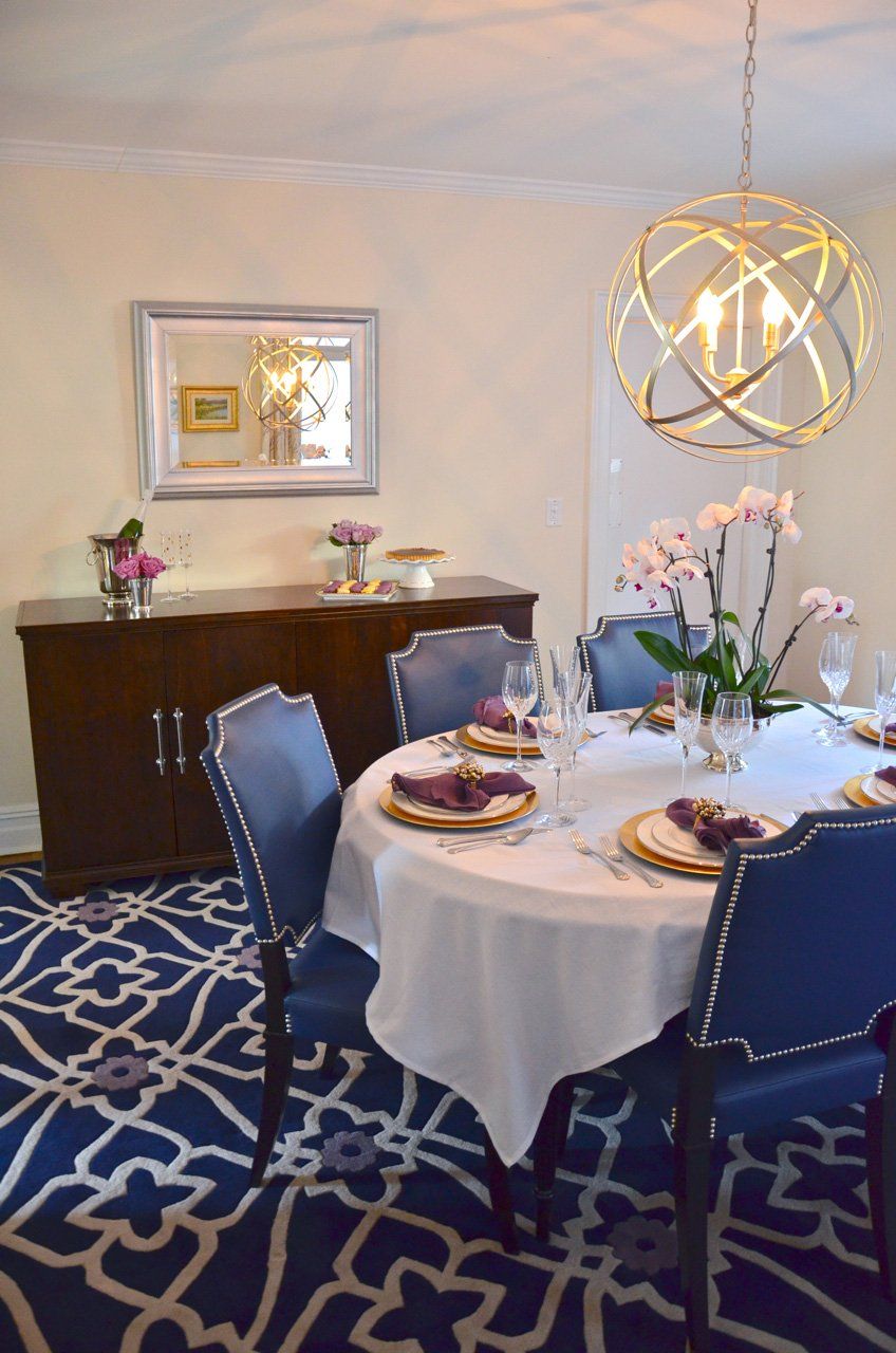 Dining room with round table set for guests, blue chairs, buffet, and patterned rug.