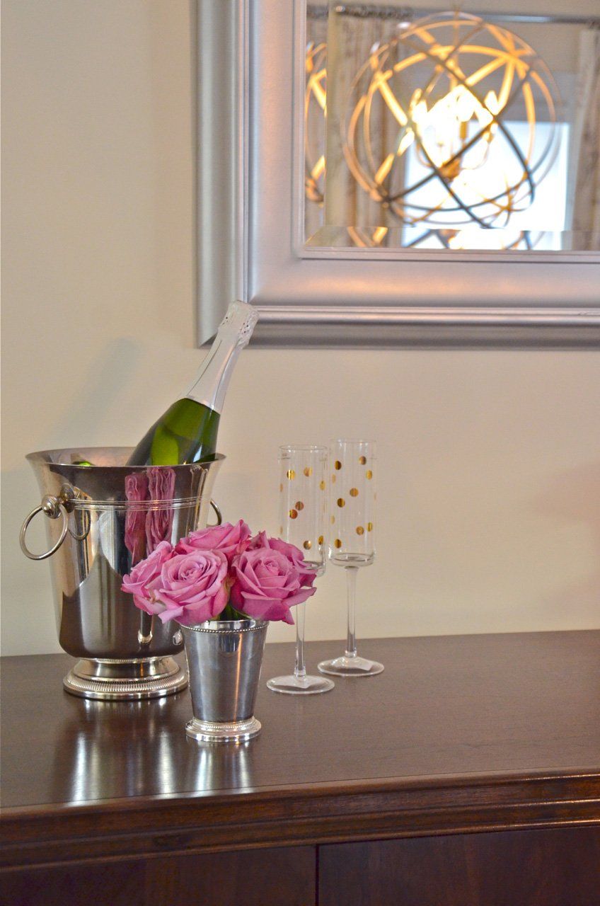 Champagne bottle in ice bucket, two flutes, and roses on a dark wood surface; mirror with light fixture in background.