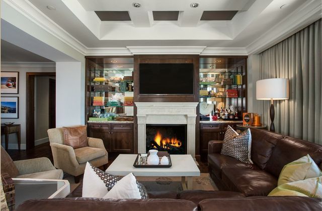 Living room with fireplace, built-in shelves, TV, leather sofa, and armchairs. Warm colors, cozy setting.