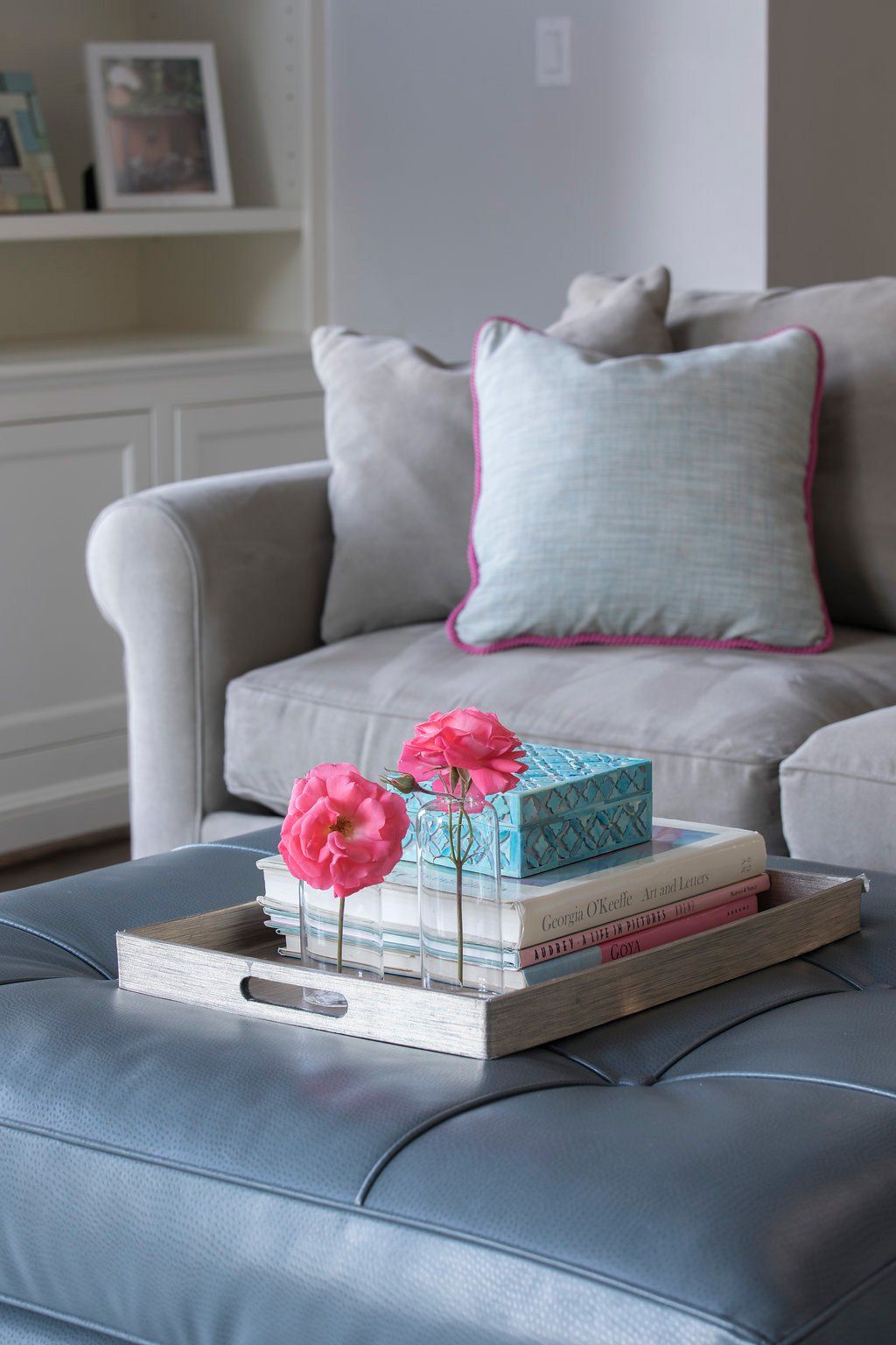 Gray sofa with light blue and pink accent pillow, ottoman with tray holding flowers and books.