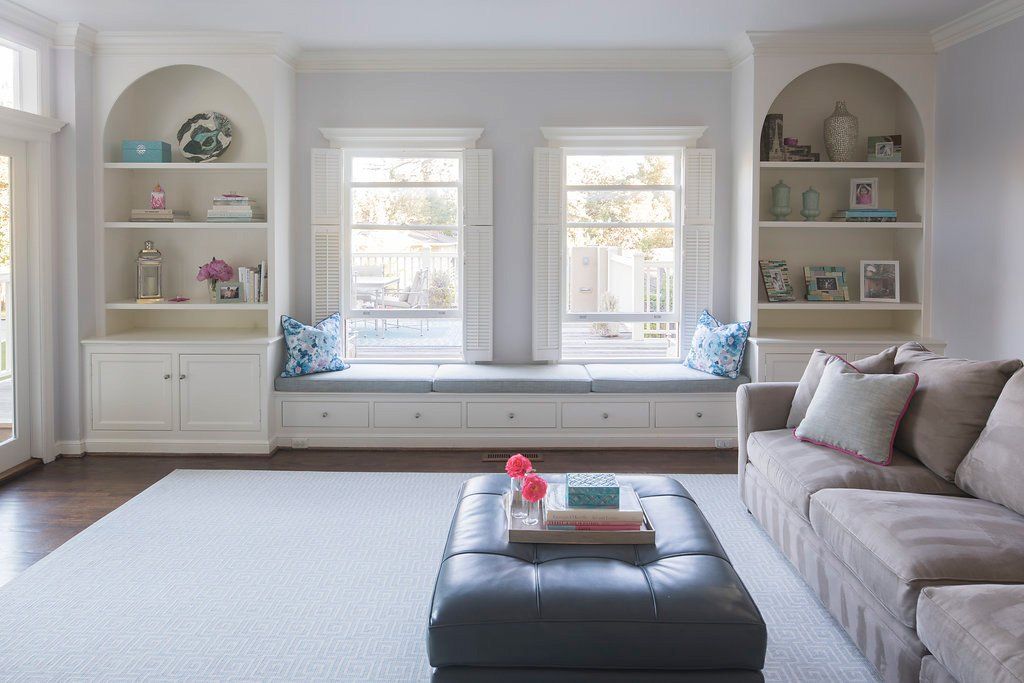 Living room with built-in bookshelves, windows with bench seating, gray couch, and a blue ottoman on a light gray rug.