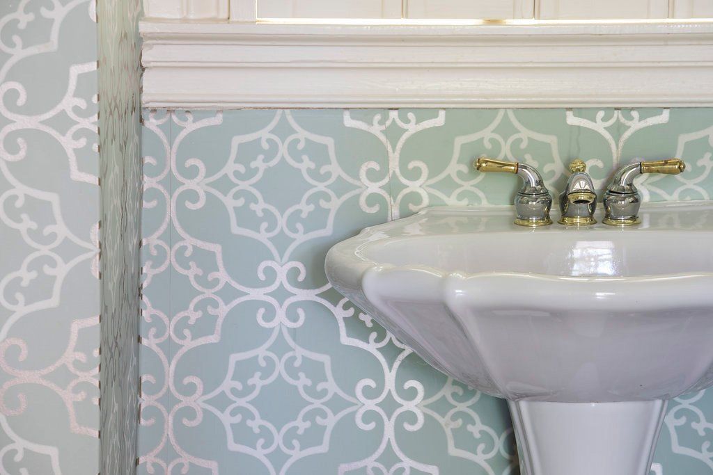 Pedestal sink with gold fixtures, against pale blue patterned wallpaper and white molding.