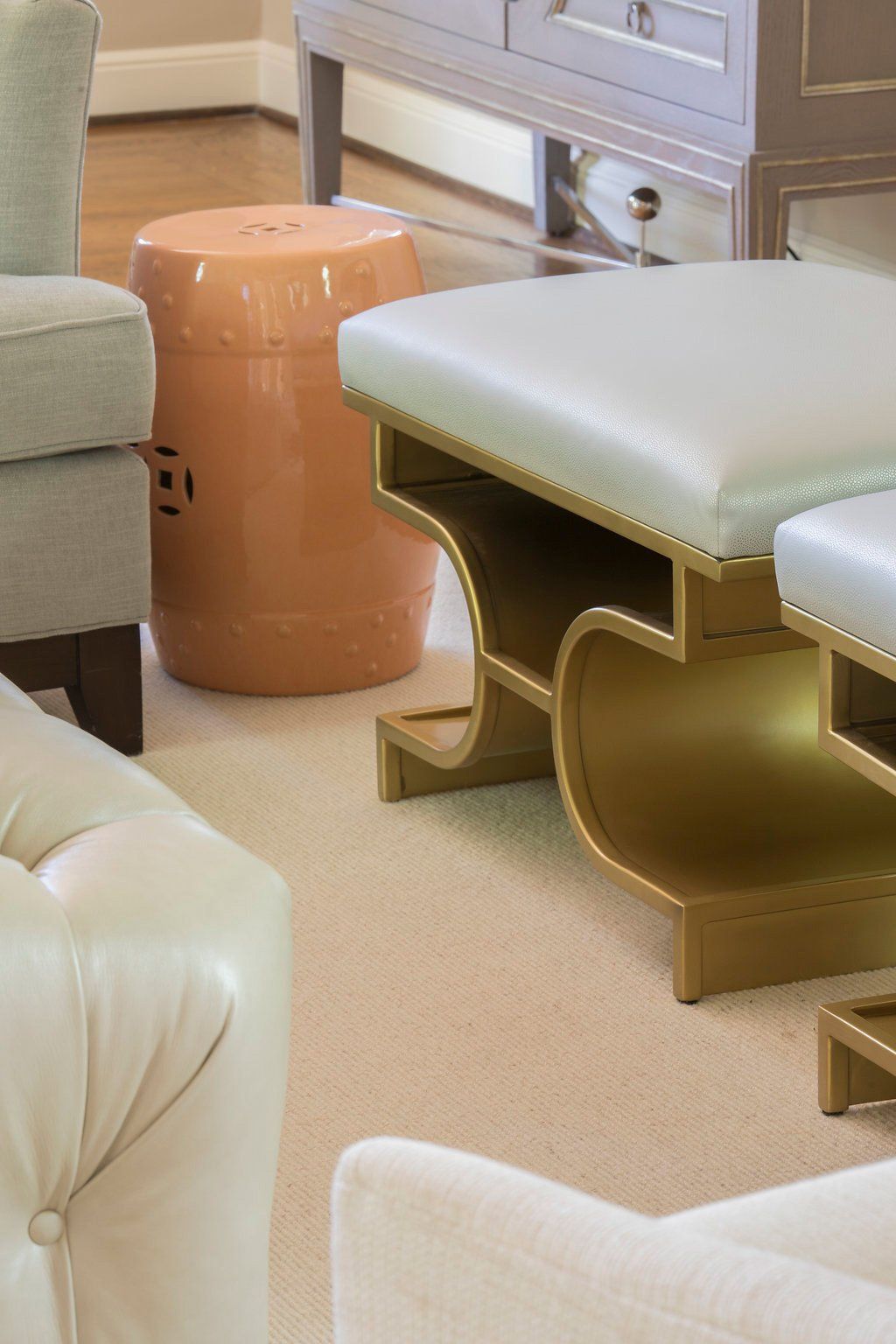 A room with various light-colored furniture, including a gold-framed ottoman with a cream-colored seat and a peach ceramic stool.