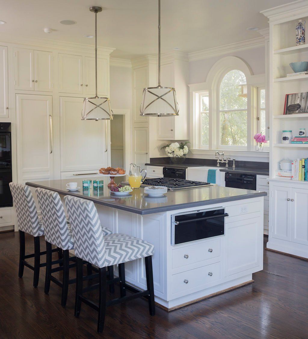 White kitchen with island, pendant lights, and patterned bar stools; dark countertop and cabinets.