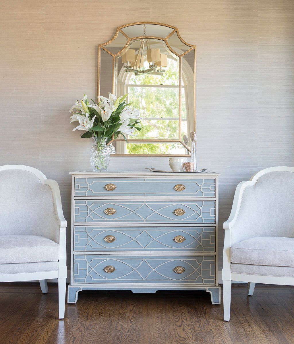 A chest of drawers with a mirror, two chairs, and a vase of flowers in a light-colored room.