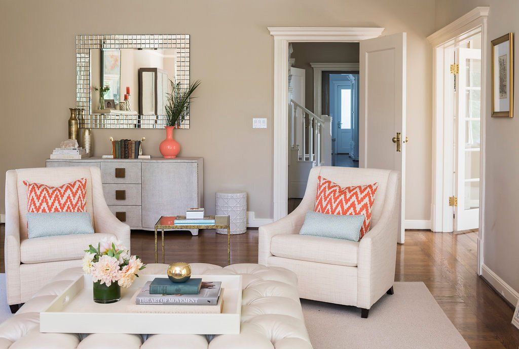 Living room with neutral tones, two armchairs with coral pillows, and a mirrored cabinet.