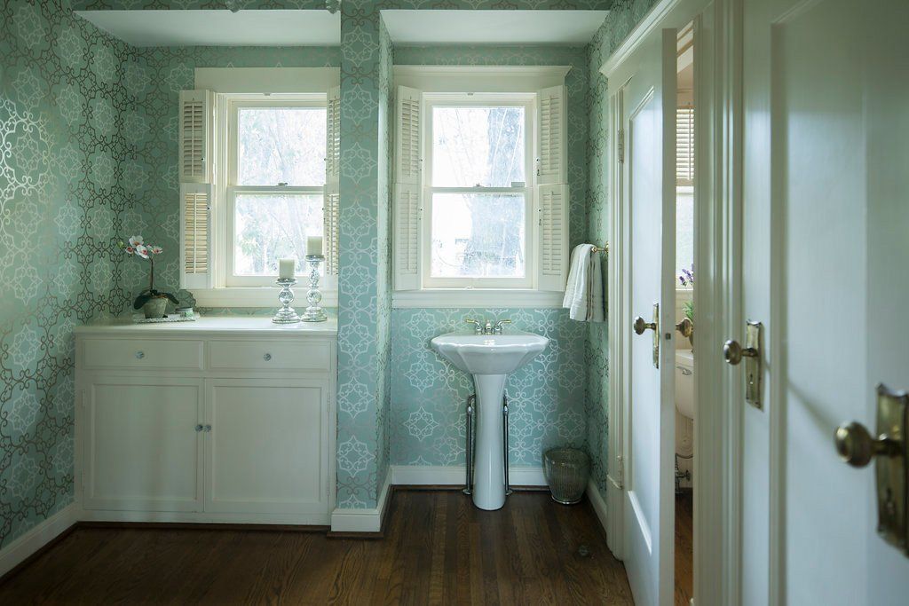 A light blue wallpapered bathroom with white cabinets, pedestal sink, shuttered windows, and a doorway.