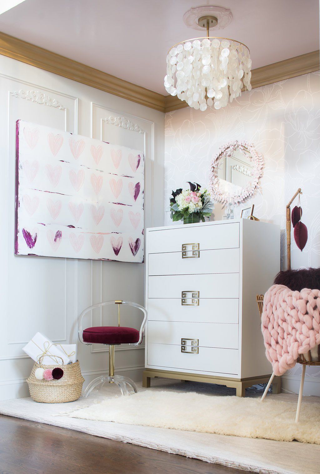 White and pink bedroom decor with chest of drawers, clear chair, art, rug, and shell chandelier.