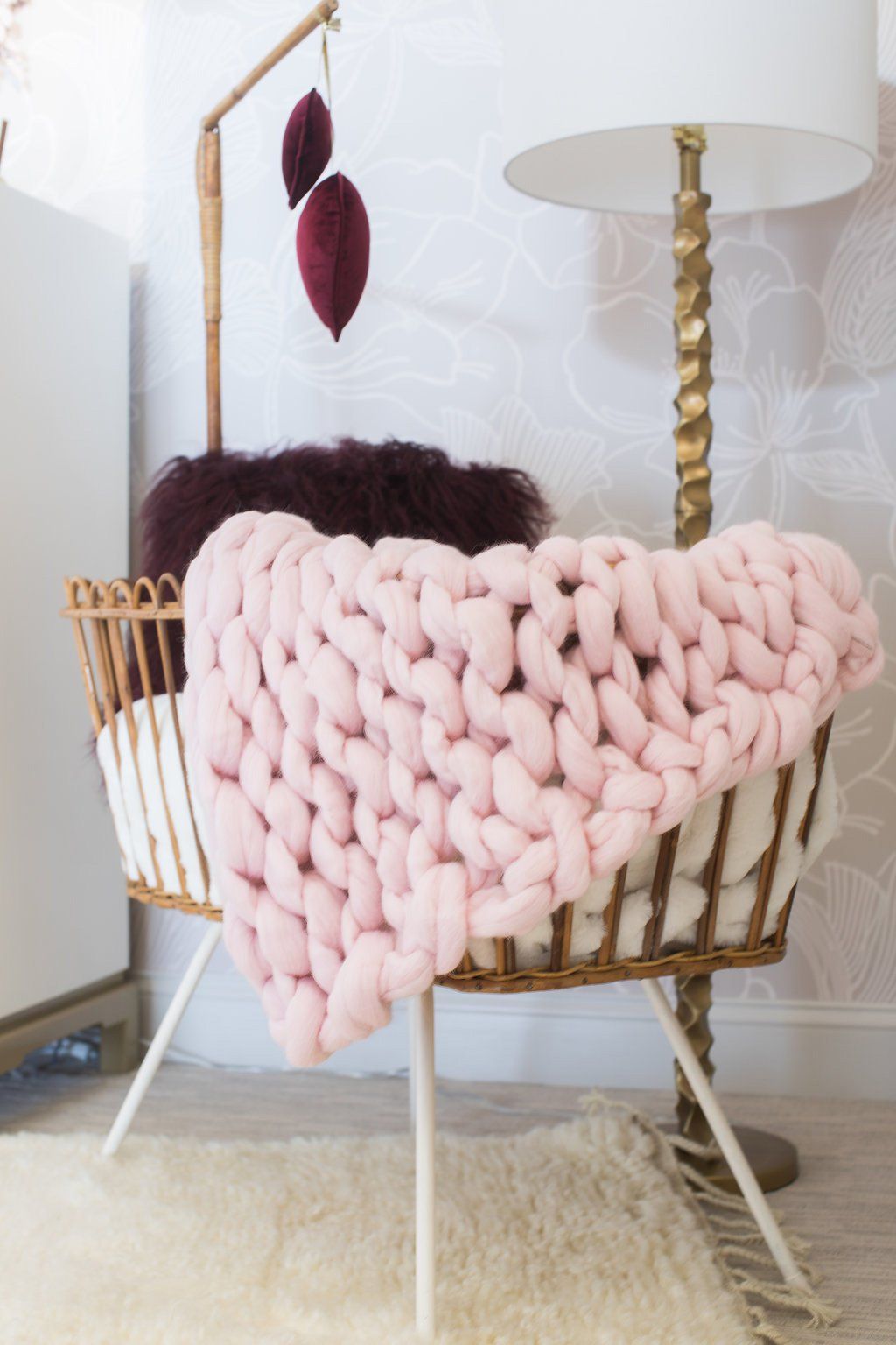 Pink chunky knit blanket draped in a wicker bassinet, next to a floor lamp and baby mobile.