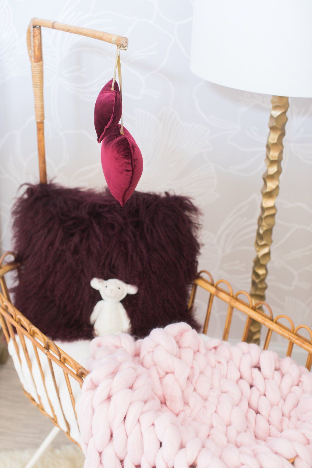 Wicker baby crib with pink blanket, fluffy burgundy pillow, white stuffed animal, and heart-shaped mobile.