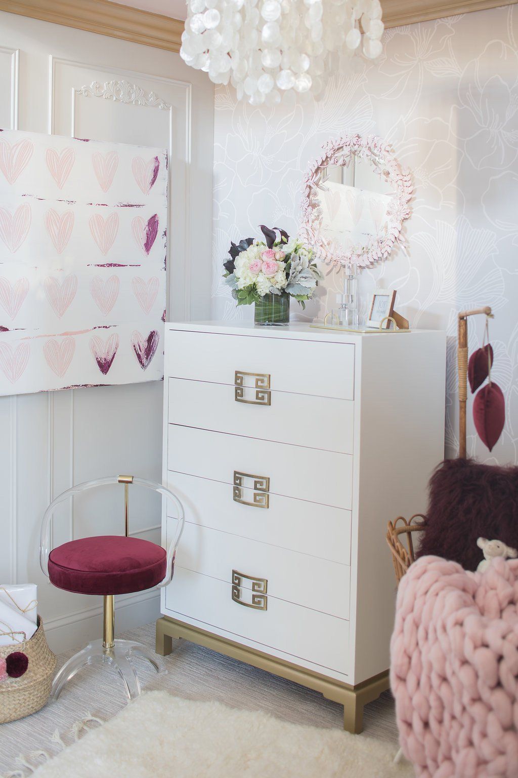 White dresser with gold accents in a bedroom, chair with red cushion, pink accents, and decorative elements.