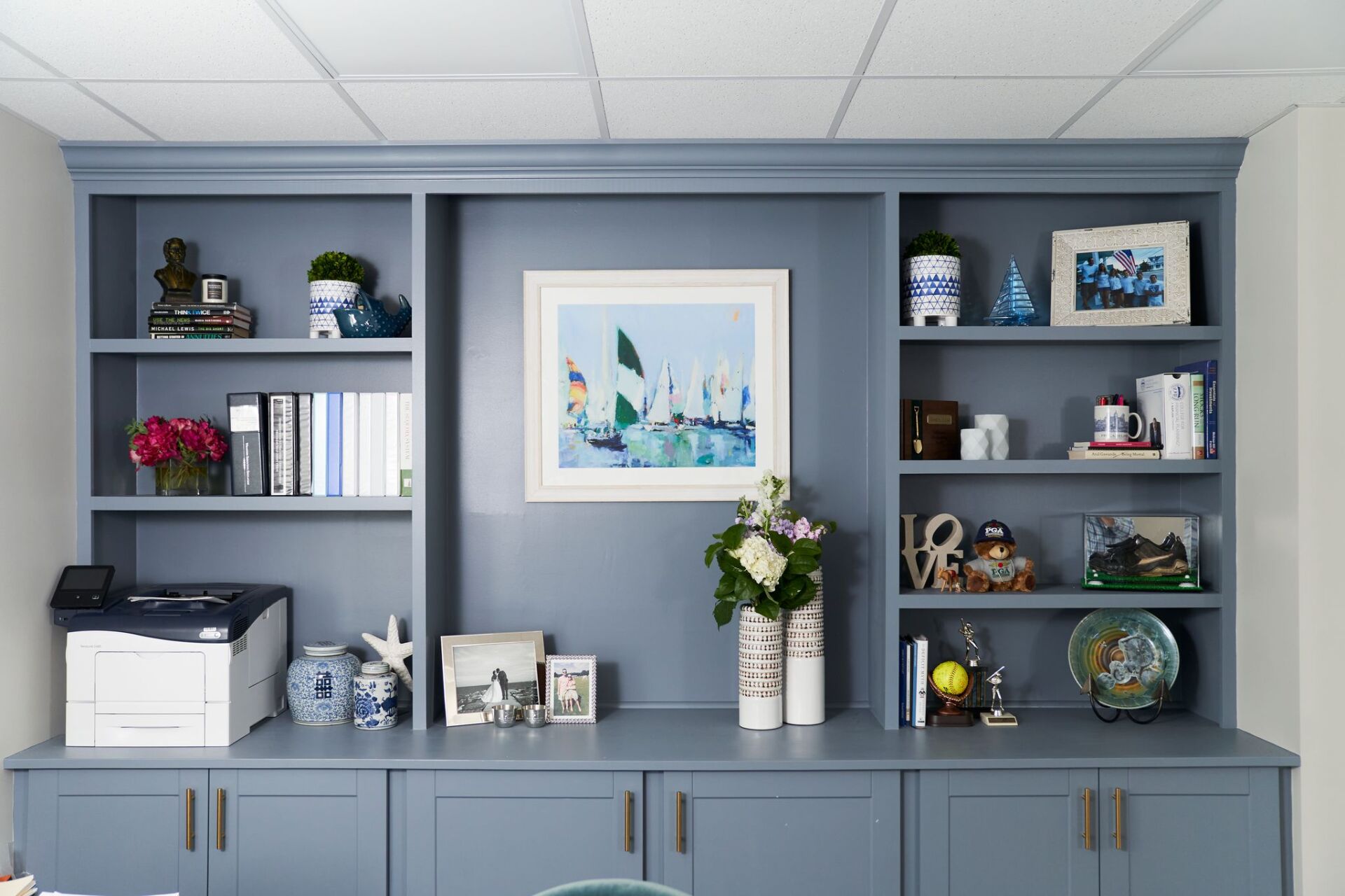 Built-in blue storage unit with shelves and cabinets, displaying books, decor, and a framed painting.