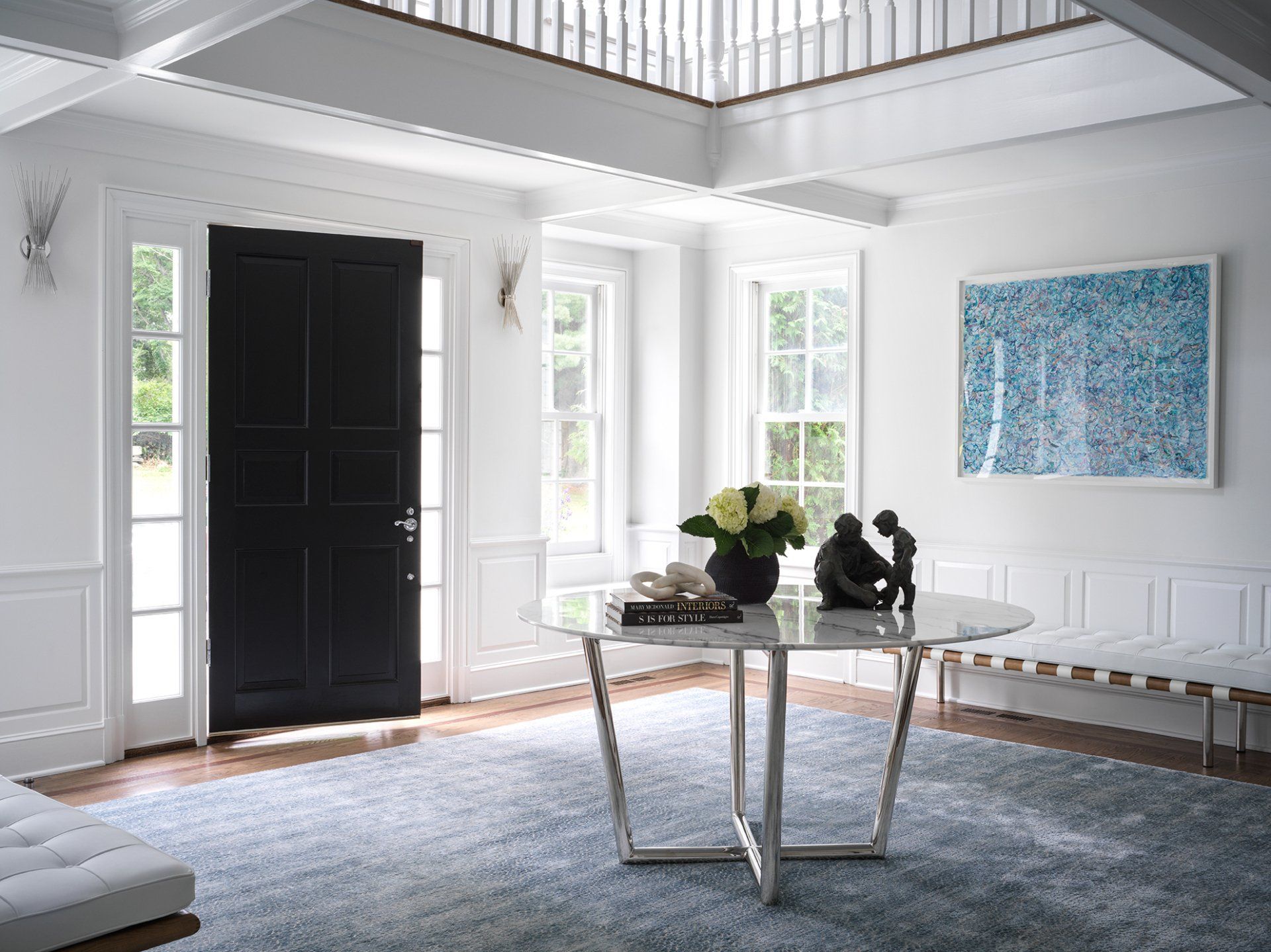 Elegant white foyer with black door, marble table, blue rug, artwork.