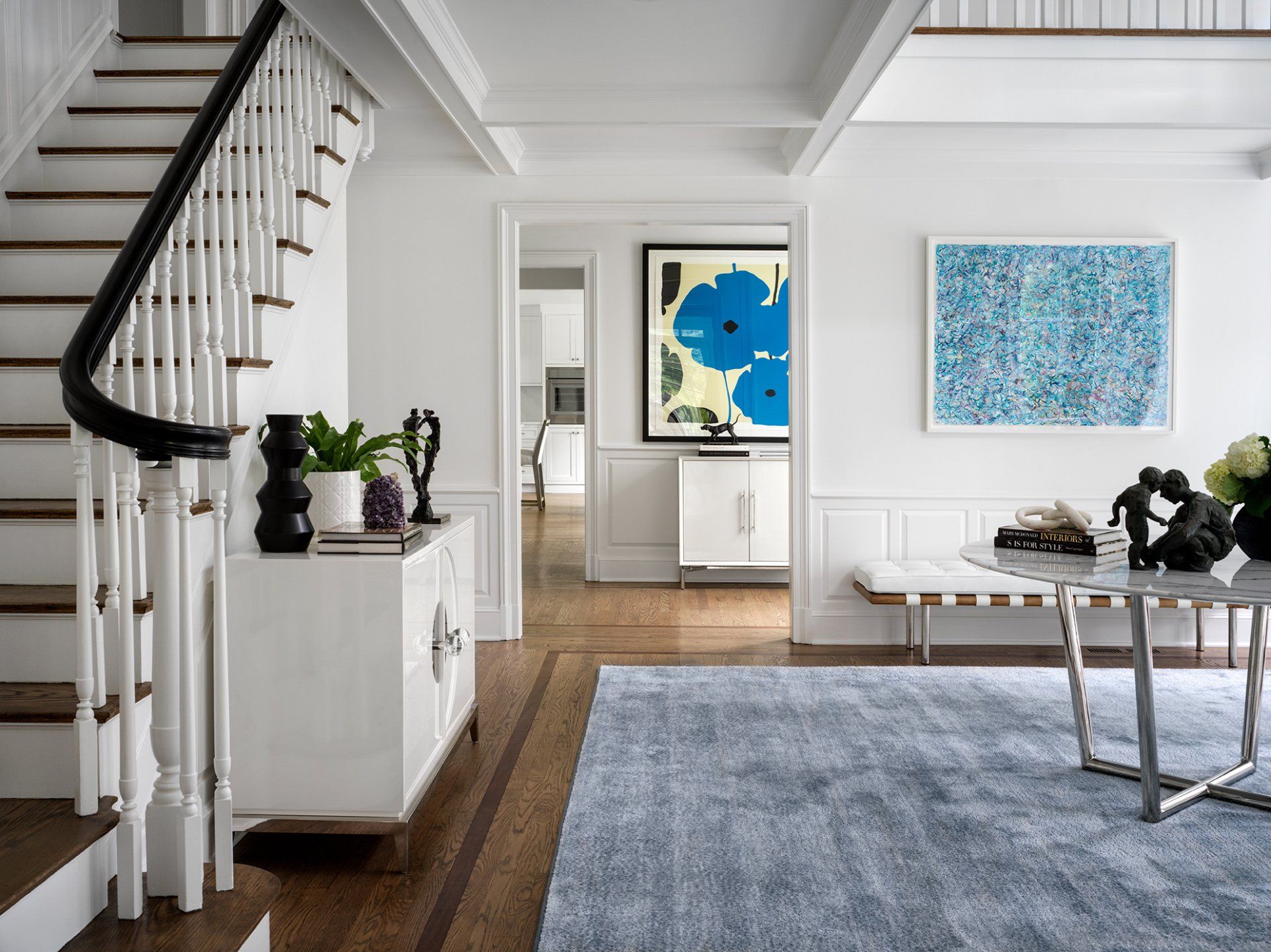 Elegant white entry hall with wood staircase, art, and light blue rug.