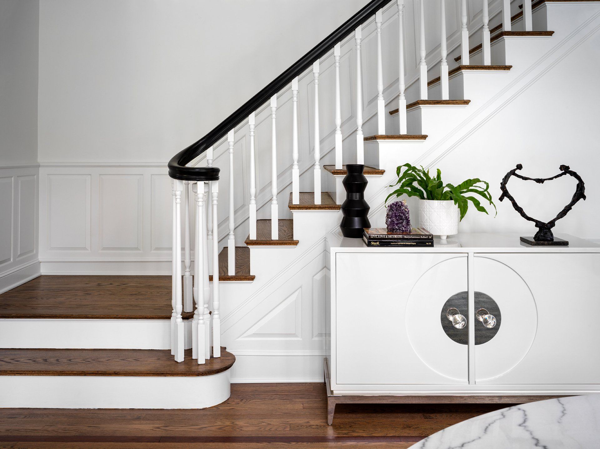 White cabinet with decor near a staircase. Black and white railing. Brown wood stairs and floor.