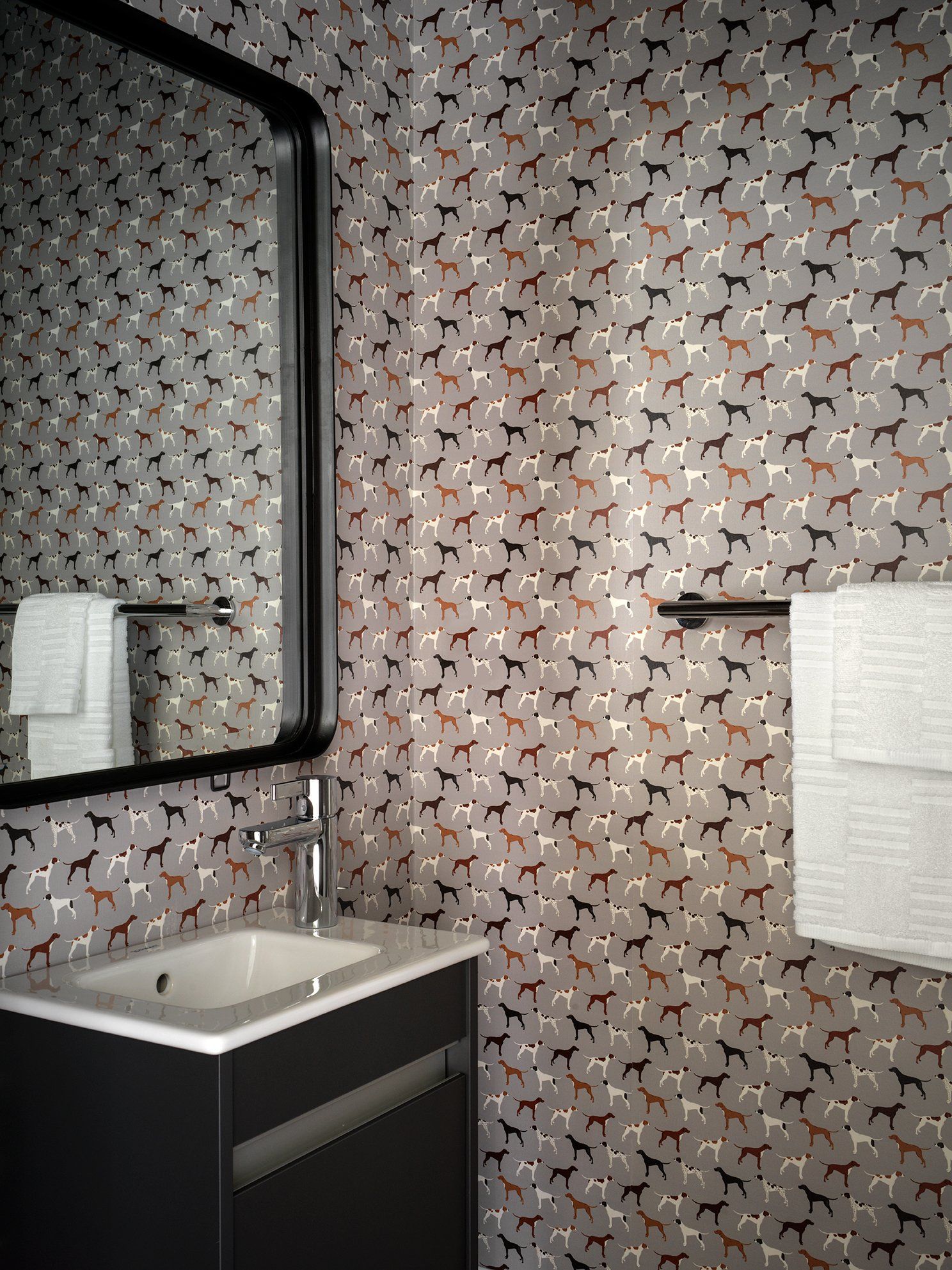 Bathroom with dog-print wallpaper, black framed mirror, gray towels, dark cabinet, and white sink.