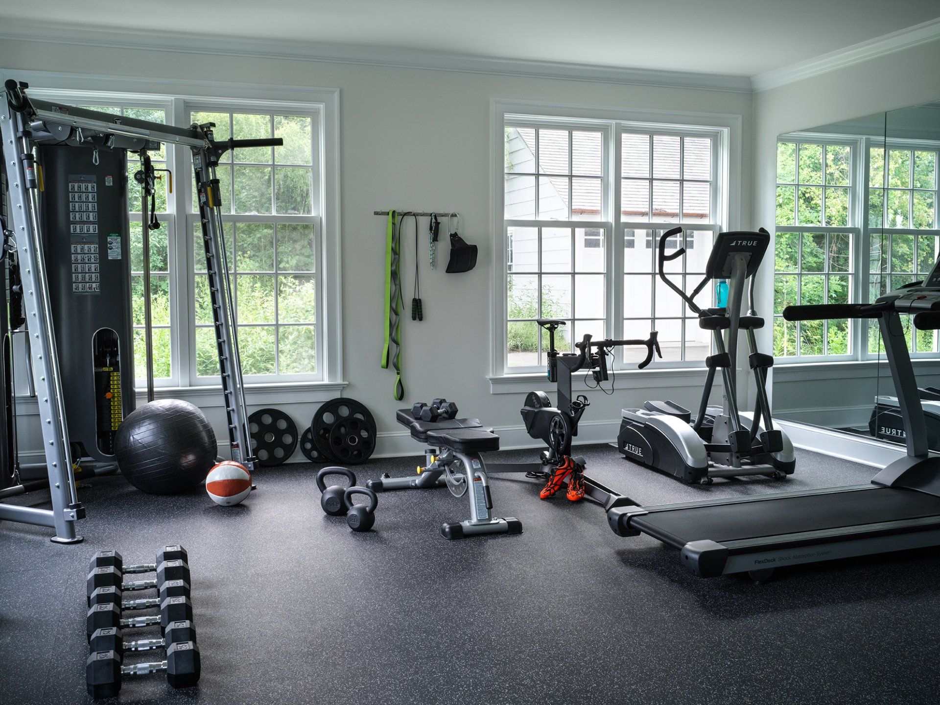 Home gym with exercise equipment: weights, elliptical, treadmill, multi-station machine, and windows.