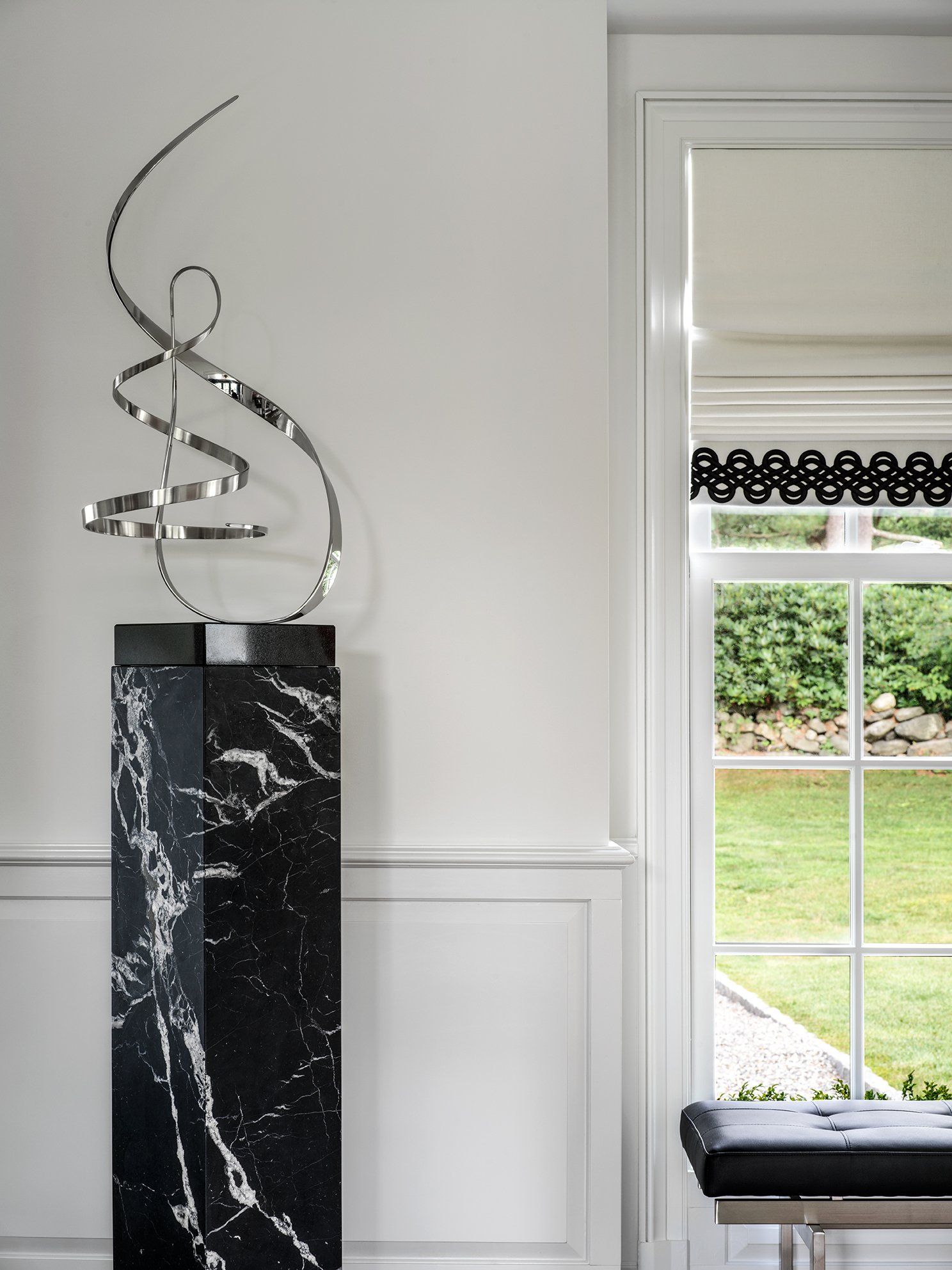 Abstract silver sculpture on a black marble pedestal, next to a window with a green view.
