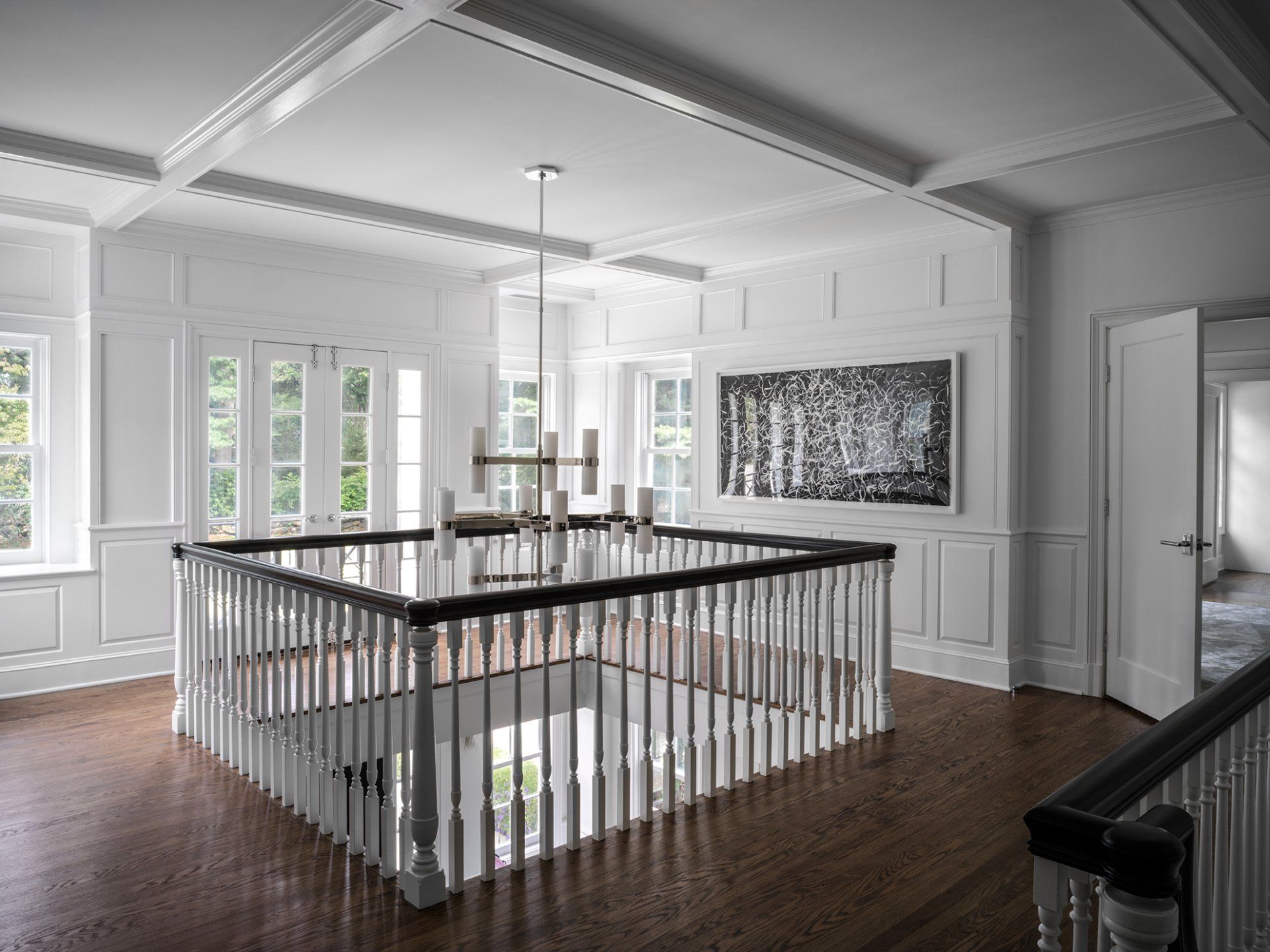 White interior with balcony, chandelier, and modern art. Light-filled space, hardwood floor, and paneled walls.