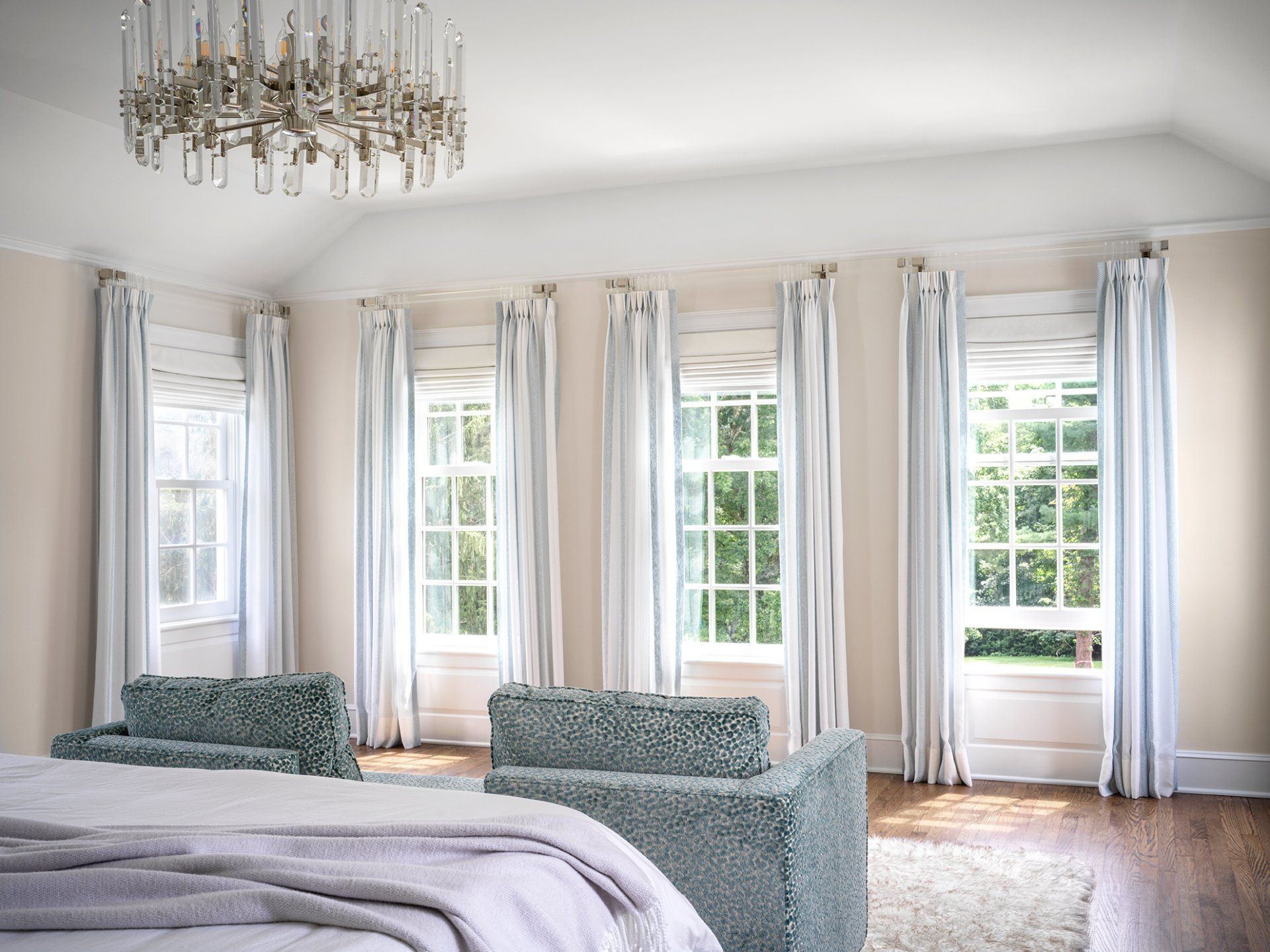 Bedroom with four windows, white curtains, blue chairs, and a chandelier.
