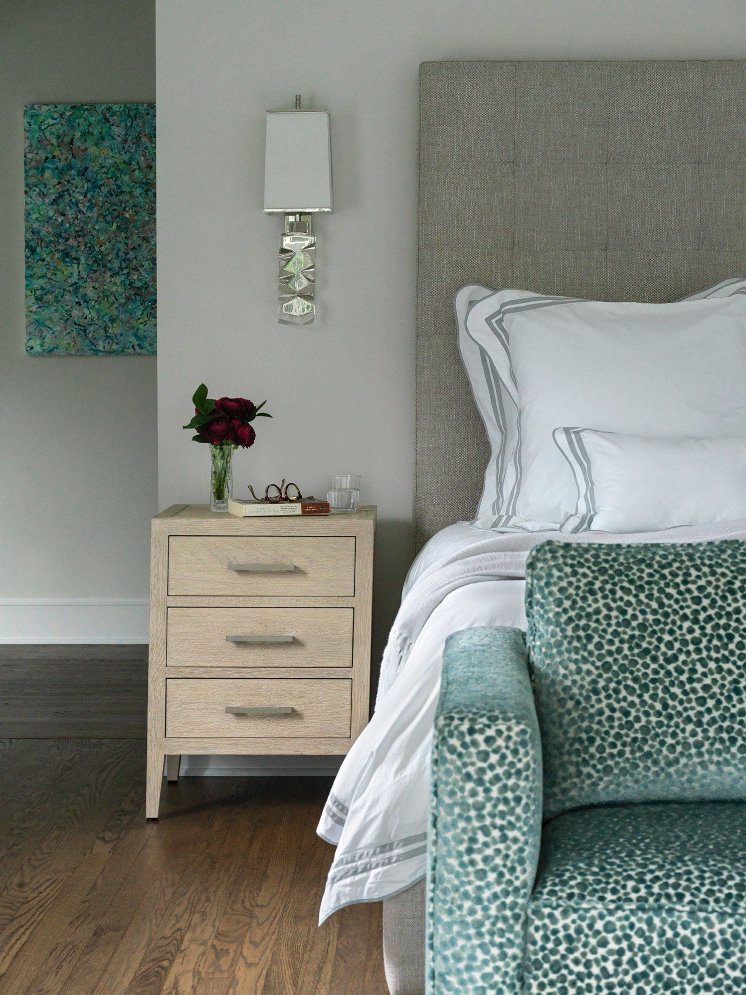 Bedroom with a textured headboard, white bedding, teal accent chair, and a nightstand with a wall-mounted light.