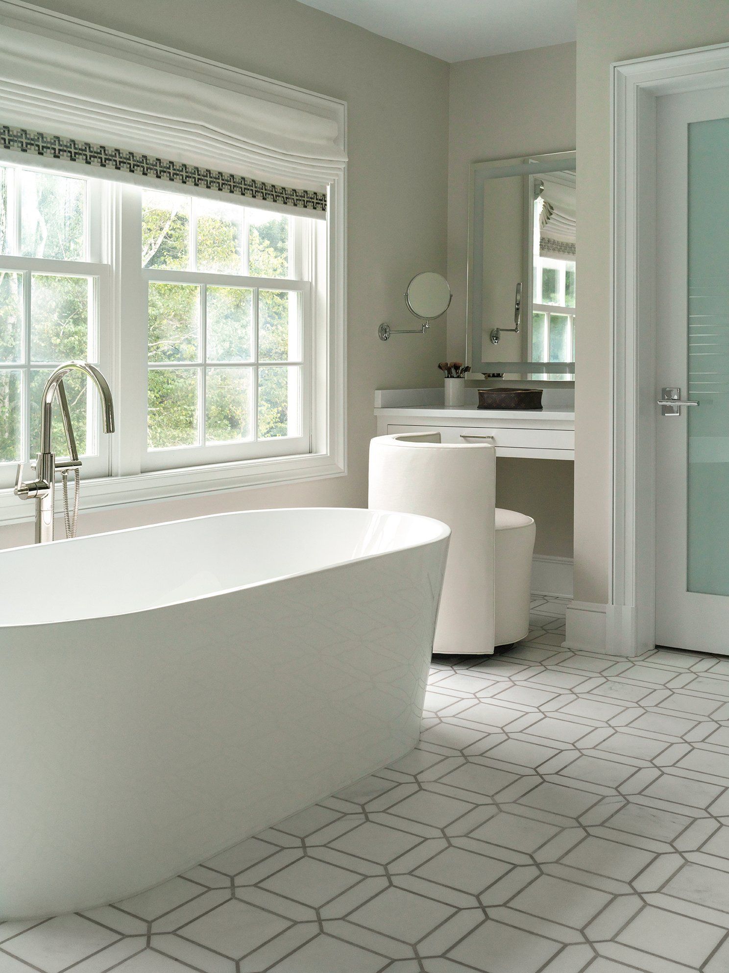 White, modern bathroom with a soaking tub by a window and geometric floor tiles.