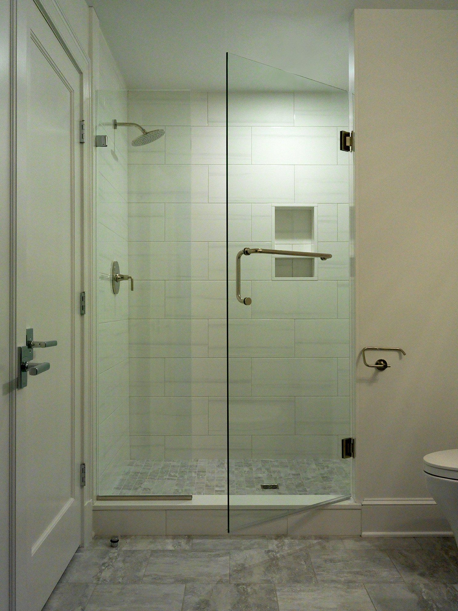 Glass-walled shower with stone floor and white tiled walls. Silver fixtures, including showerhead and door handle, are visible.