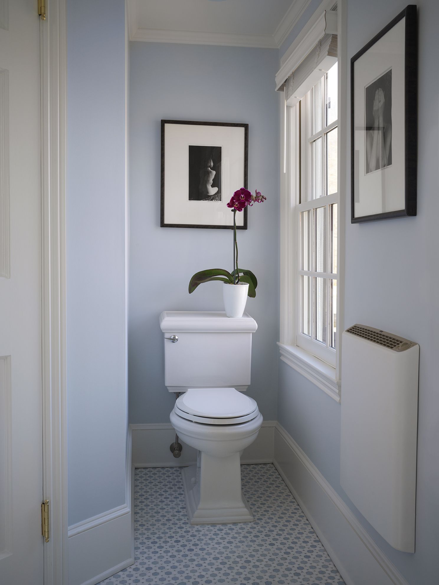 Small powder room with blue walls, white toilet, and patterned floor tiles. Framed art and a window with a shade.
