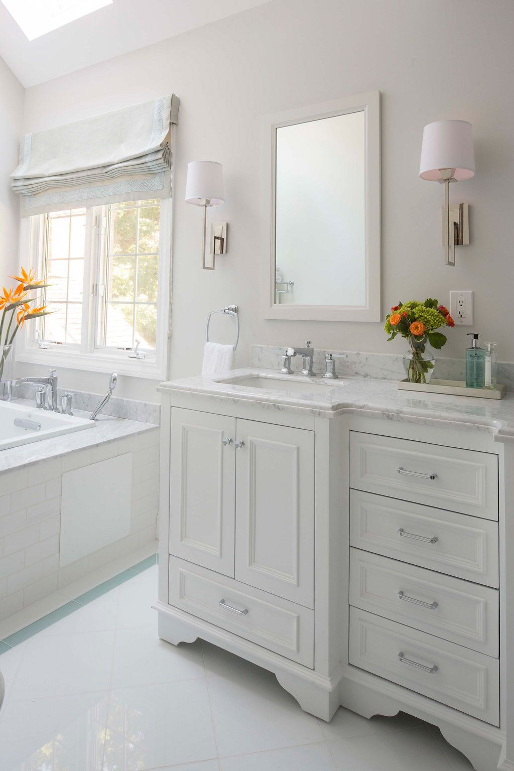 White bathroom with vanity, mirror, and sconces. Bathtub near window with Roman shade.