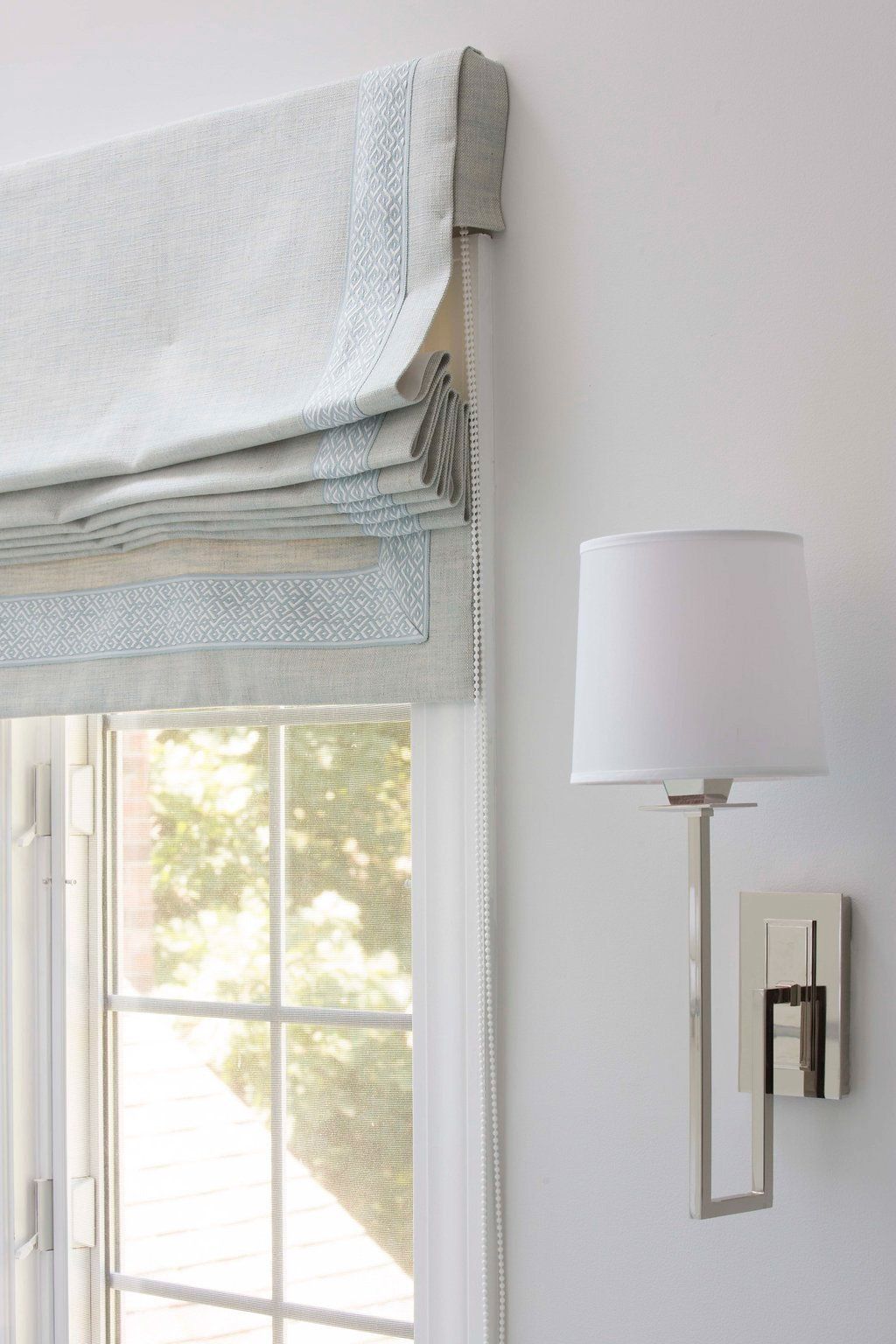 Light blue Roman shade above a window, next to a silver wall sconce.