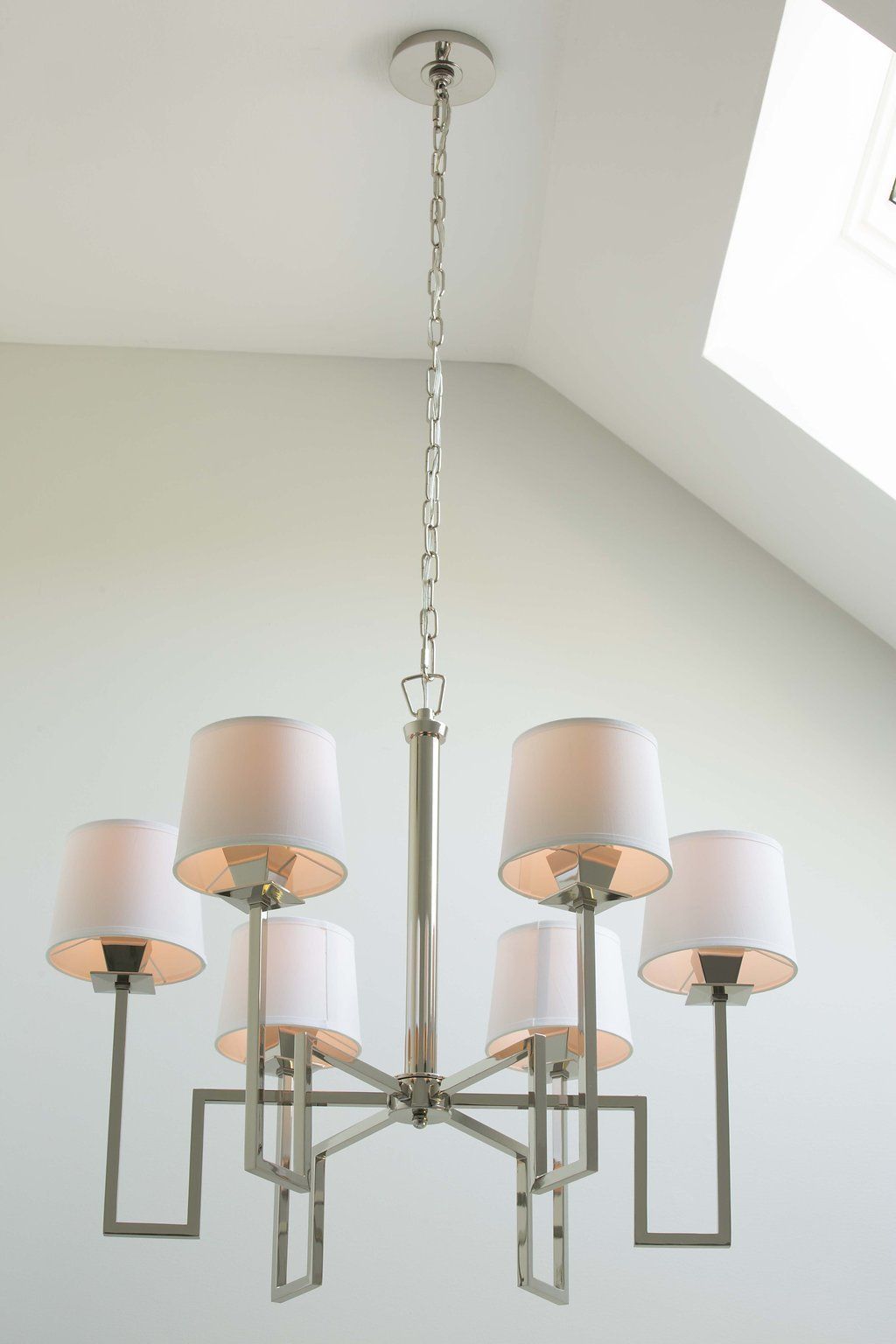 Silver chandelier with six white lampshades suspended from a chain in a room with a skylight.