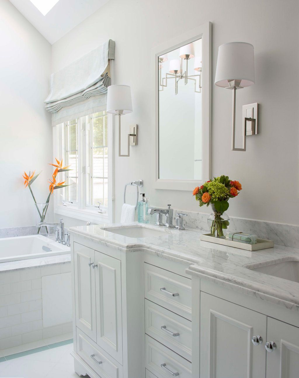 White bathroom with double vanity, bathtub, large mirror, and floral arrangements.
