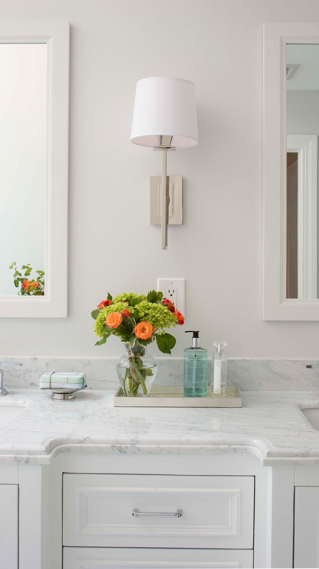 Bathroom vanity with white marble countertop, flowers, and a wall lamp.
