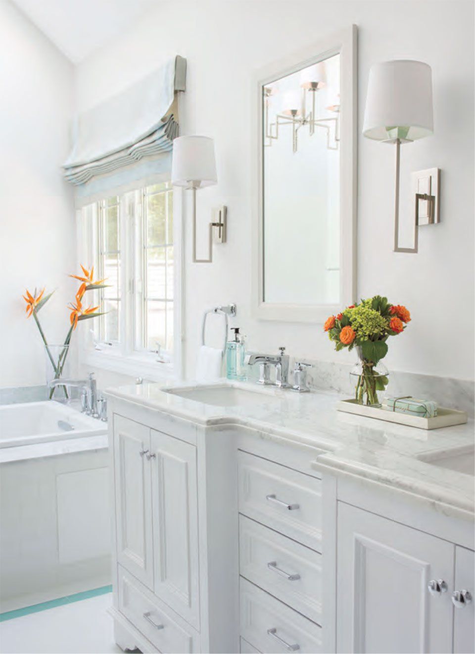 White bathroom with vanity, marble countertop, mirror, sconces, window with blue shade, and flowers.