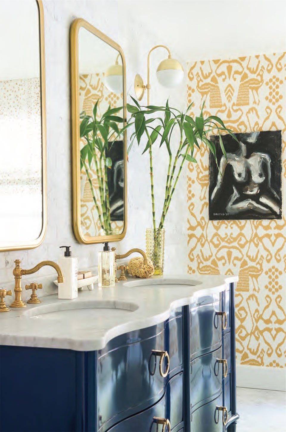 Bathroom with a blue vanity, gold fixtures, and yellow patterned wallpaper.  A painting hangs on the wall, and mirrors reflect bamboo.