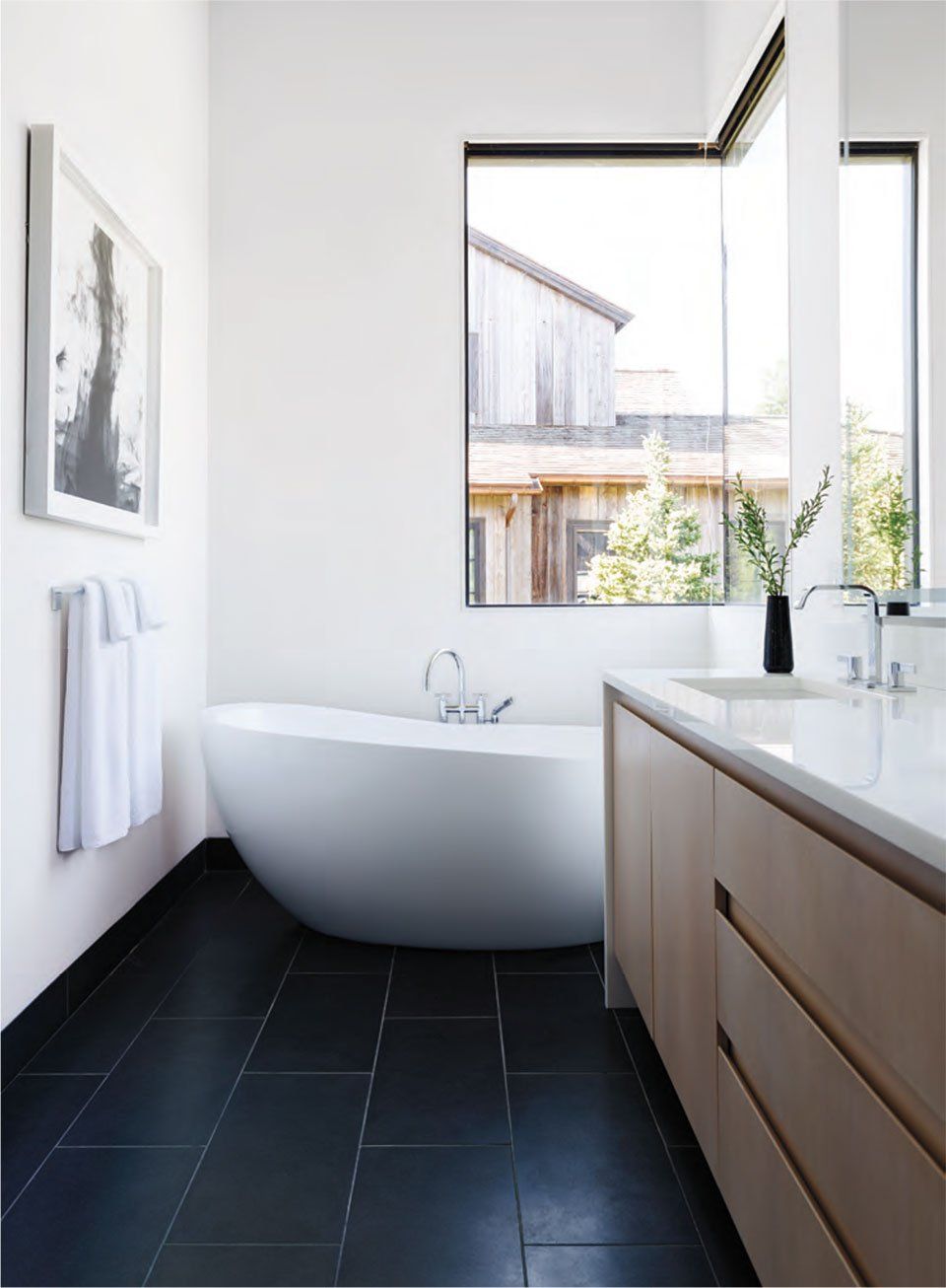 Modern bathroom with a white oval tub, black tile floor, and large window.
