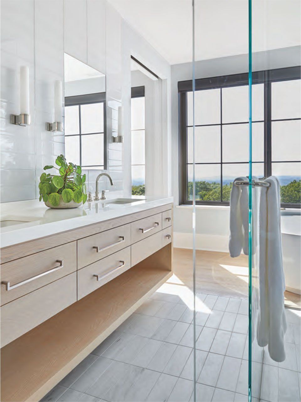 Modern bathroom with light wood vanity, glass shower, large windows.