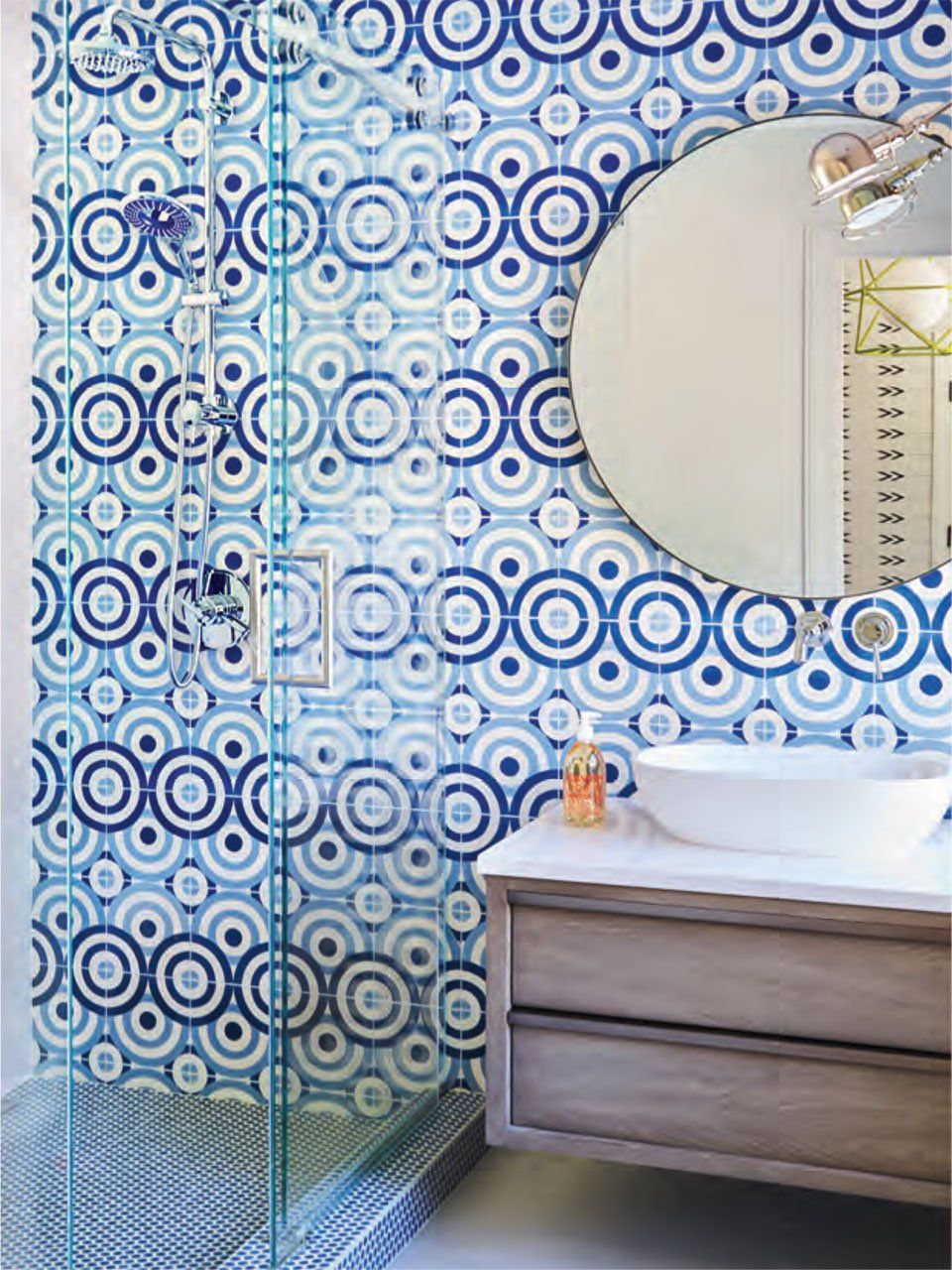 Blue and white patterned tile shower with a round mirror and a wooden vanity.