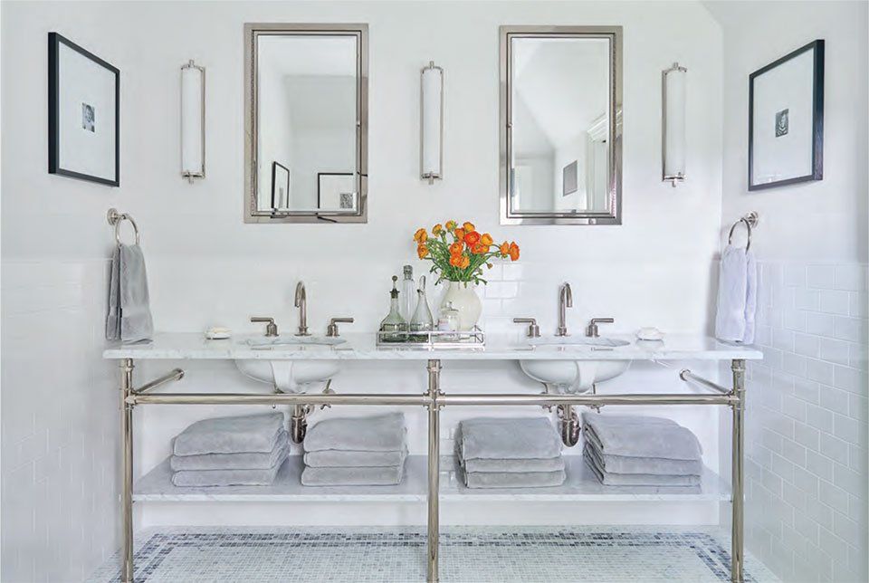 White bathroom with double sinks, mirrors, sconces, towels, and orange flowers.
