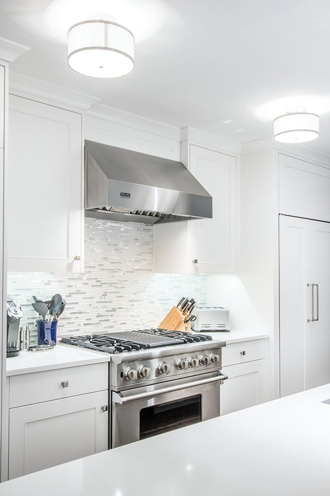 White kitchen with stainless steel range, hood, and cabinetry. Mosaic backsplash and overhead lighting.