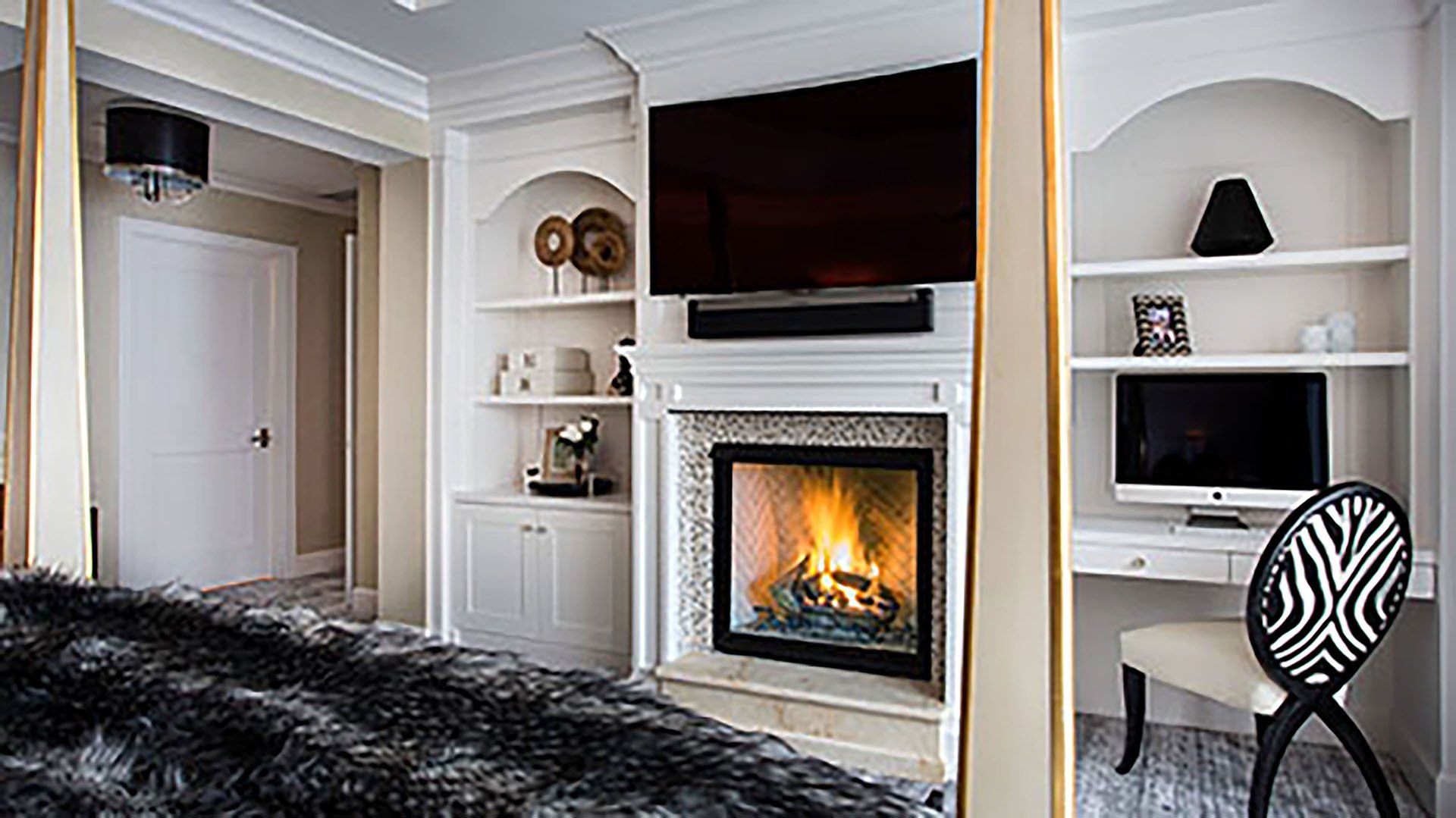 Bedroom with built-in shelves, fireplace, and TV. A zebra-print chair sits at a desk beside a bed with a fur throw.