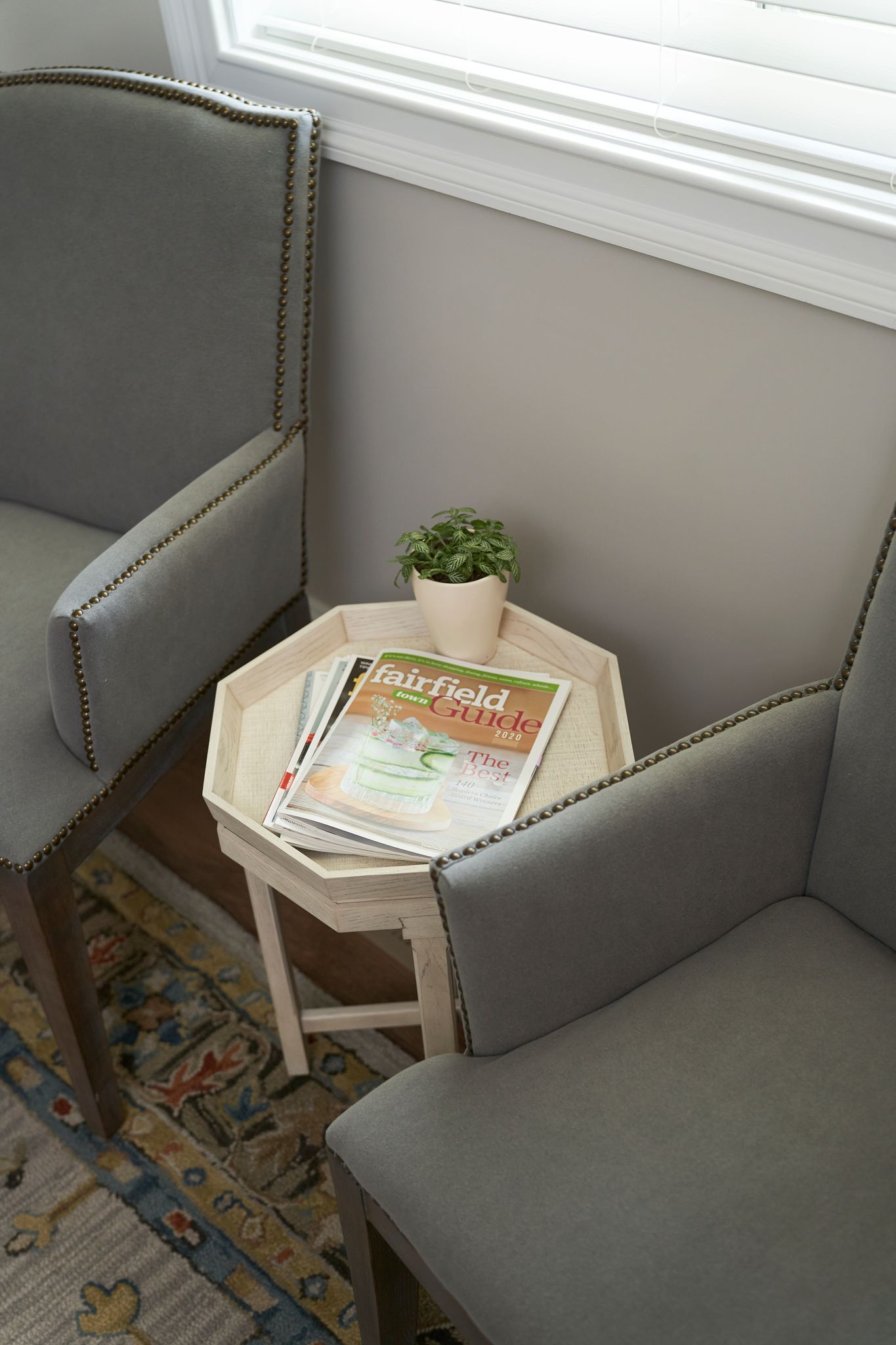 Two gray upholstered chairs flanking a small, white octagonal table with magazines and a potted plant.