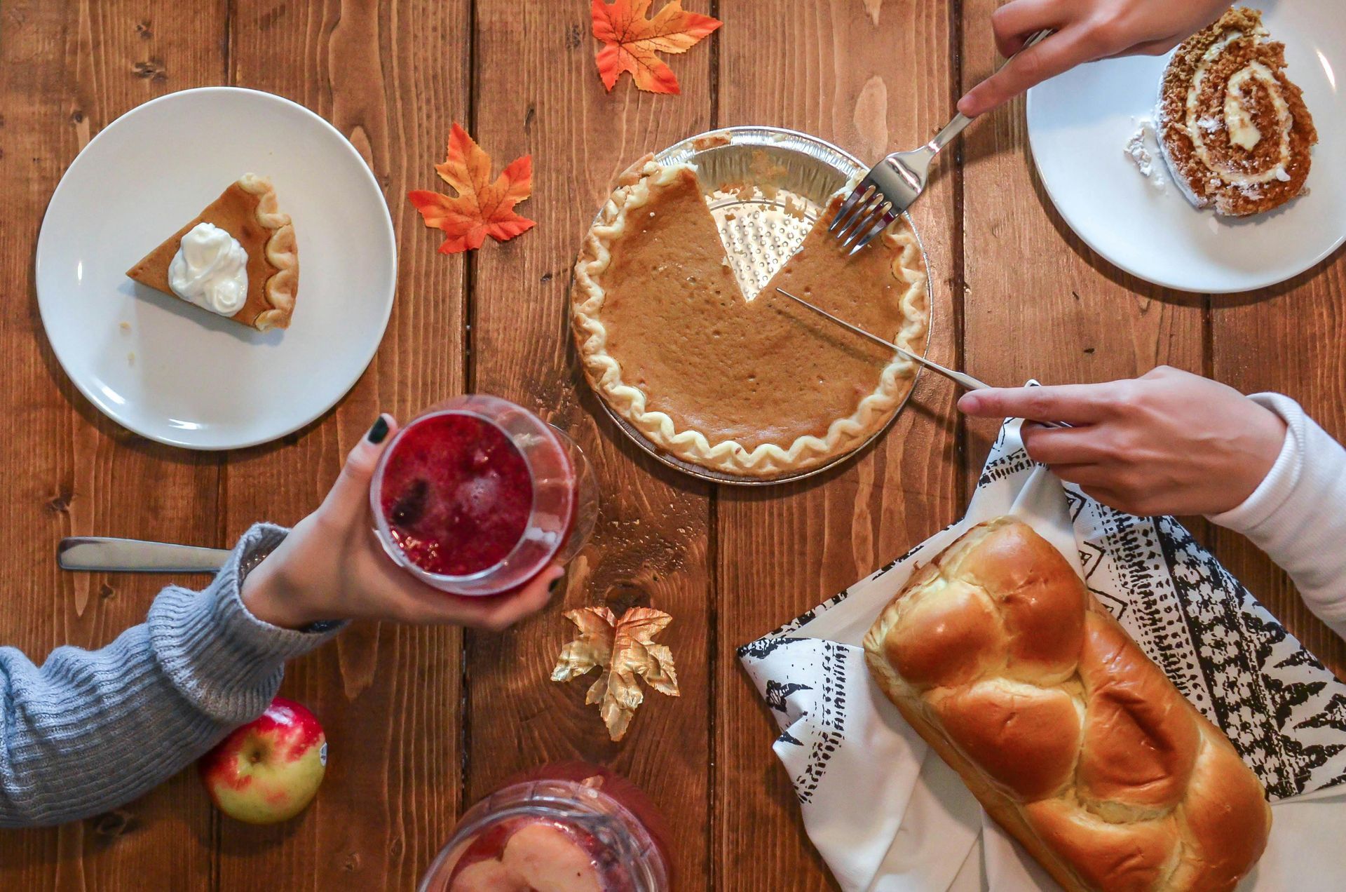 Thanksgiving dessert table: Pumpkin pie, cake, challah bread, drinks, and hands cutting pie on wooden table.