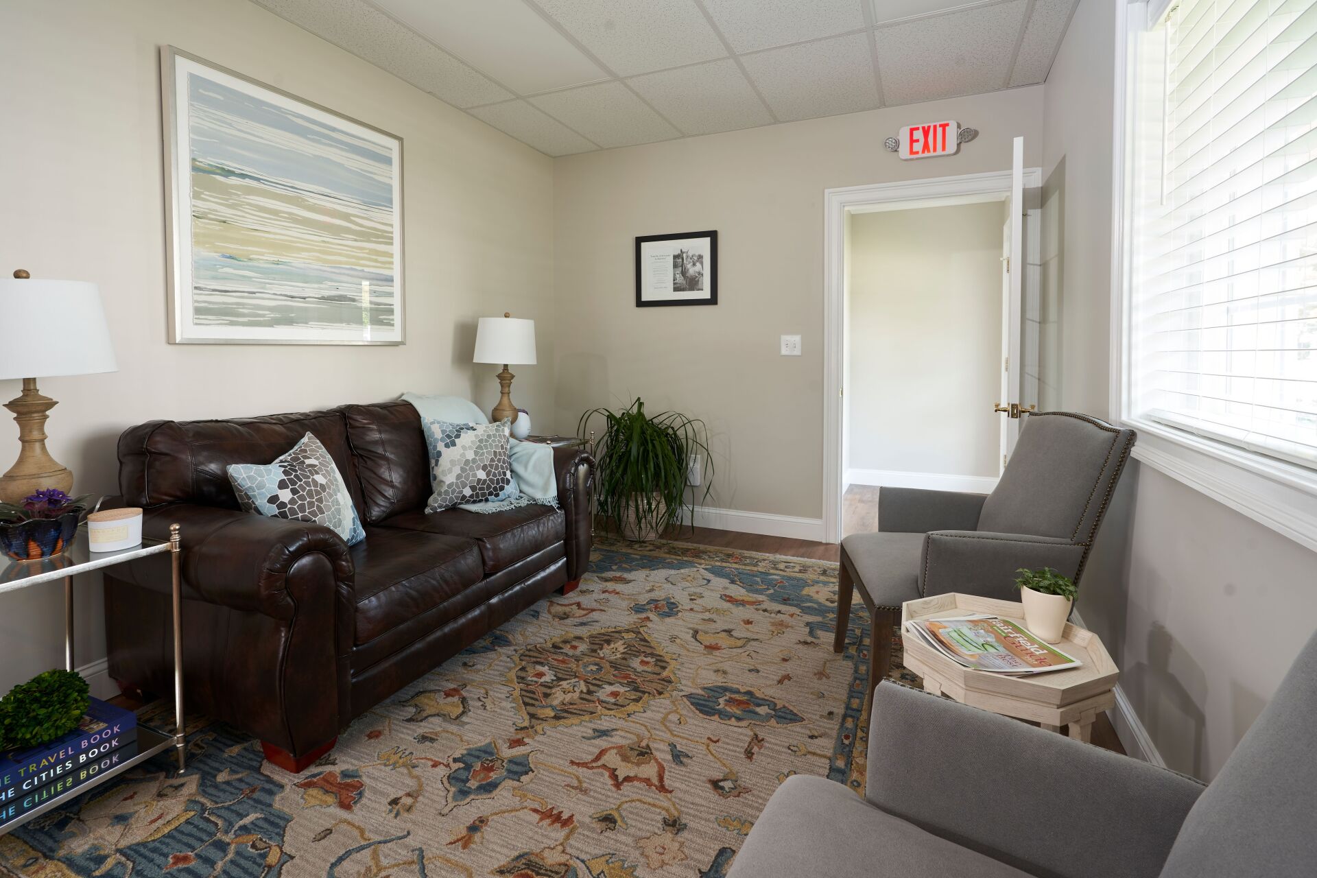 A waiting room with a brown leather couch, two gray armchairs, a patterned rug, and a framed artwork.