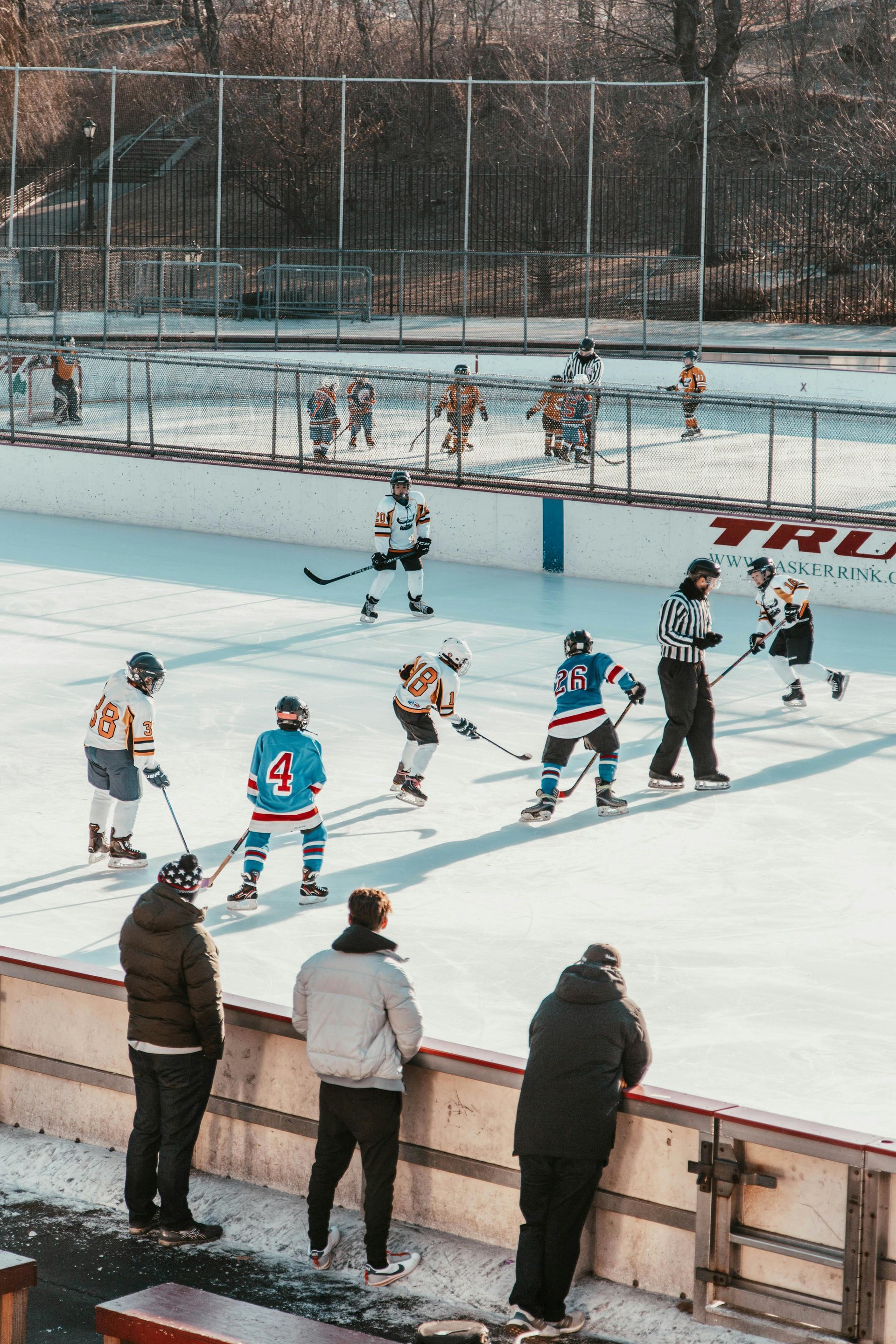 Hockey game on outdoor rink; players in jerseys skating, referee present. Spectators watch behind barrier.