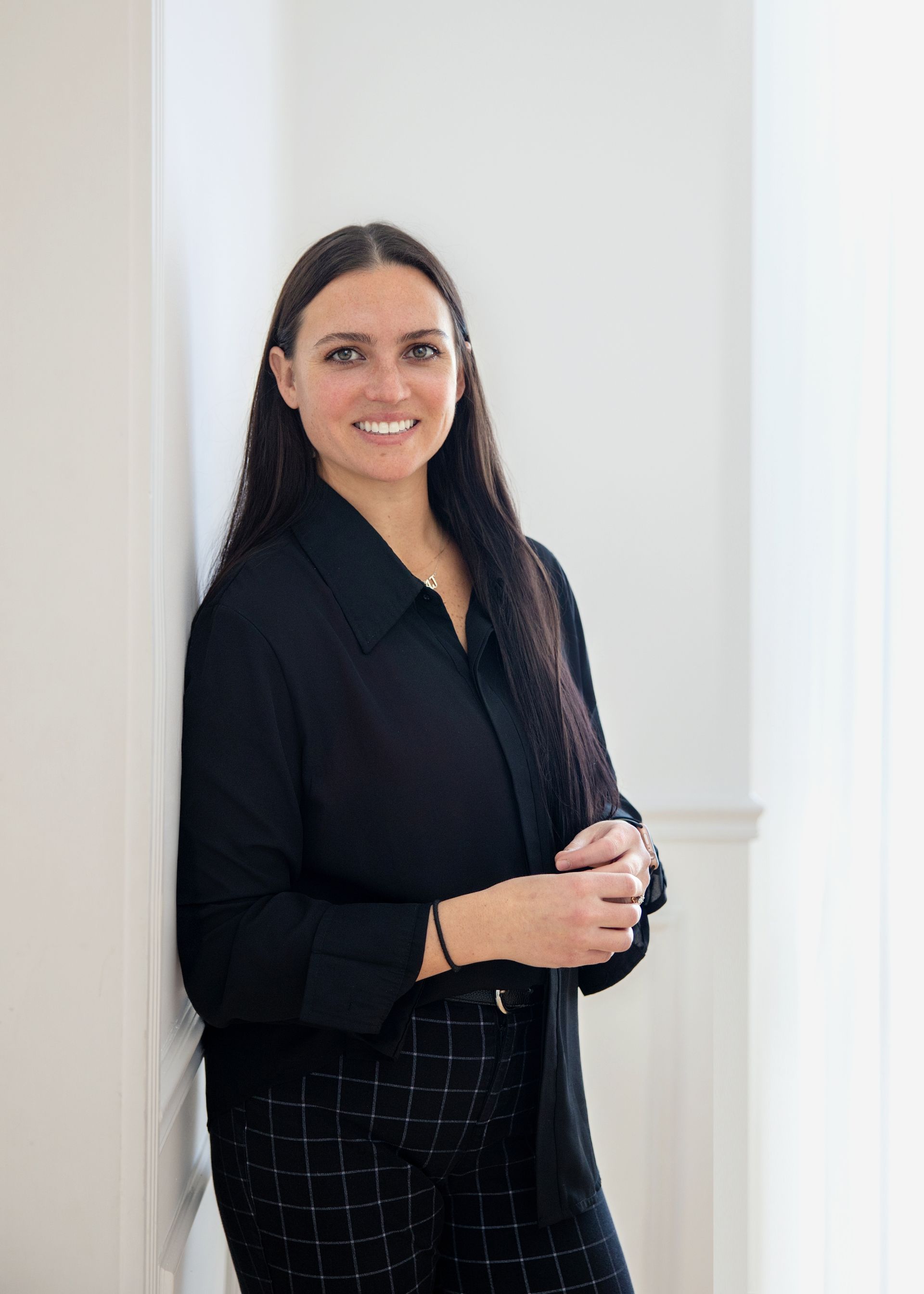Woman leaning against a white wall, smiling. Wearing a black button-down shirt and patterned pants.