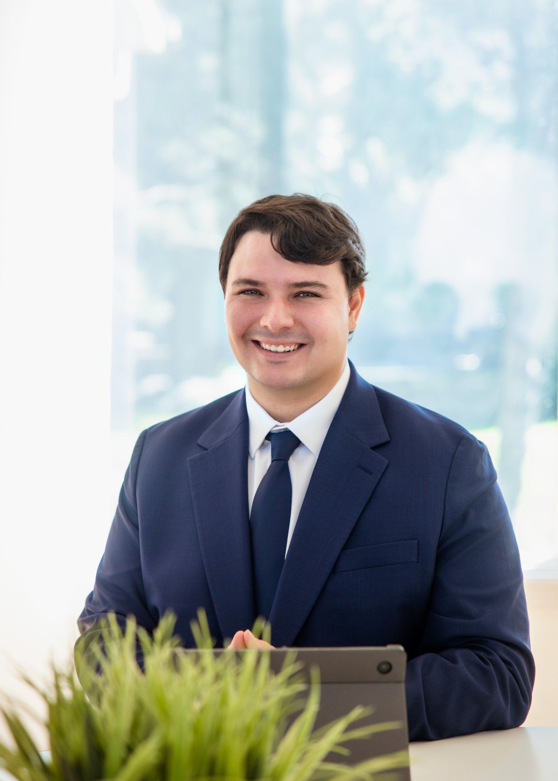 Man in a blue suit smiles, seated near a window and a plant, possibly in an office.