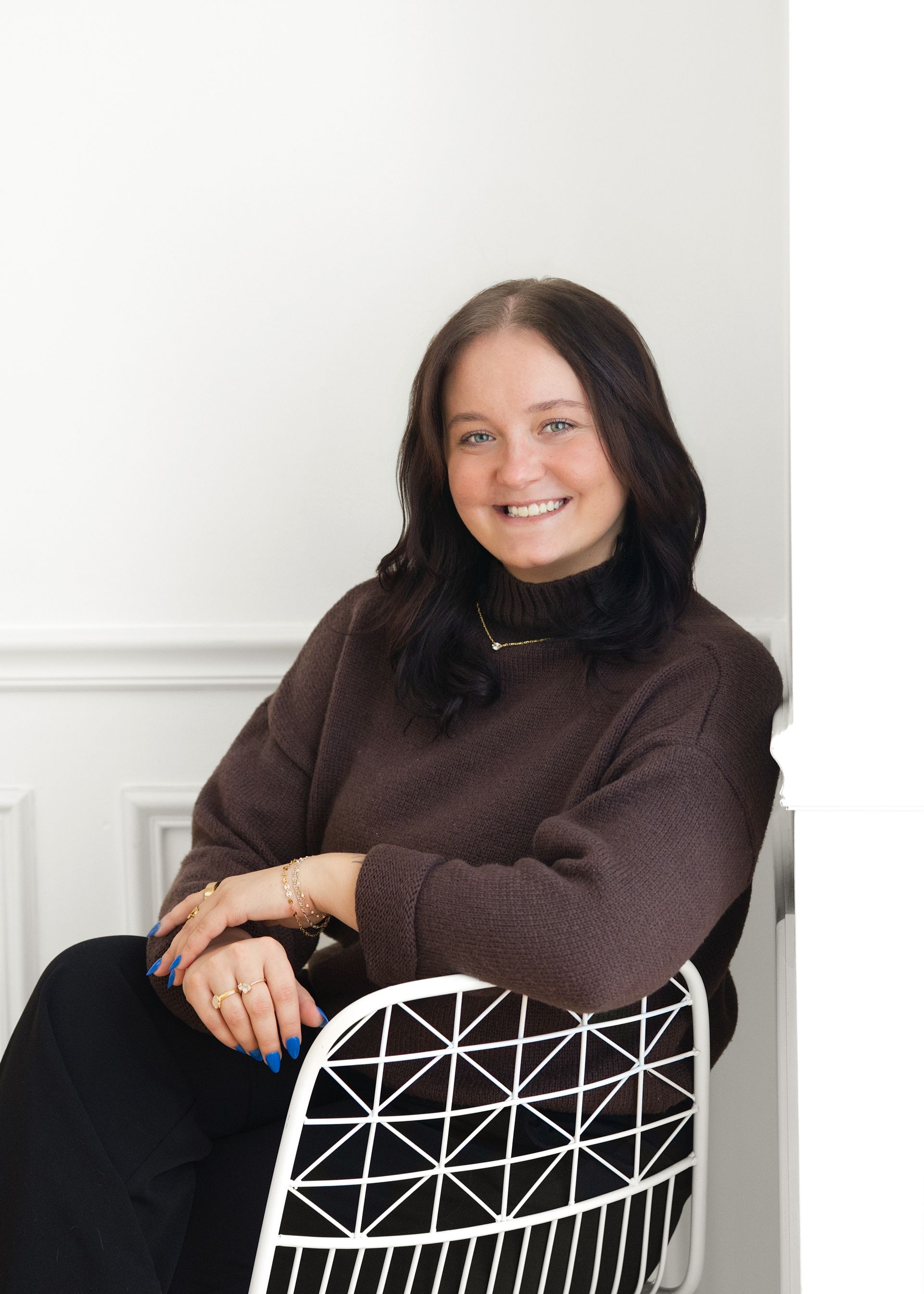 Woman in brown sweater smiles while sitting on a chair with white geometric back, leaning against a white wall.