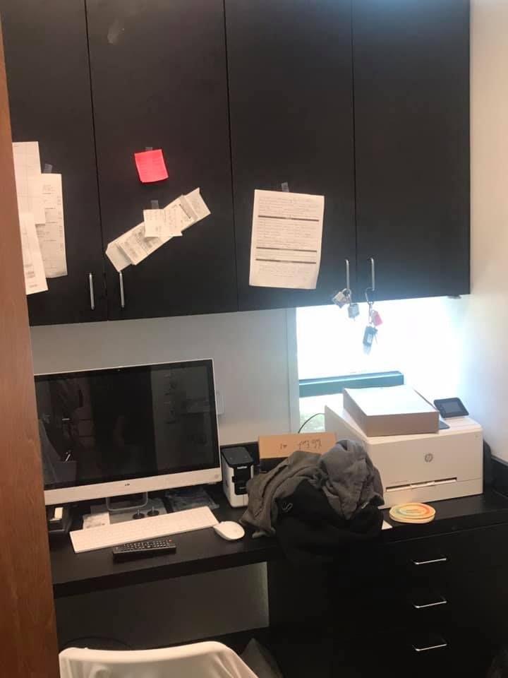 A home office with a black desk, computer, and cabinets. Papers are attached to the cabinets, and a printer sits on the desk.